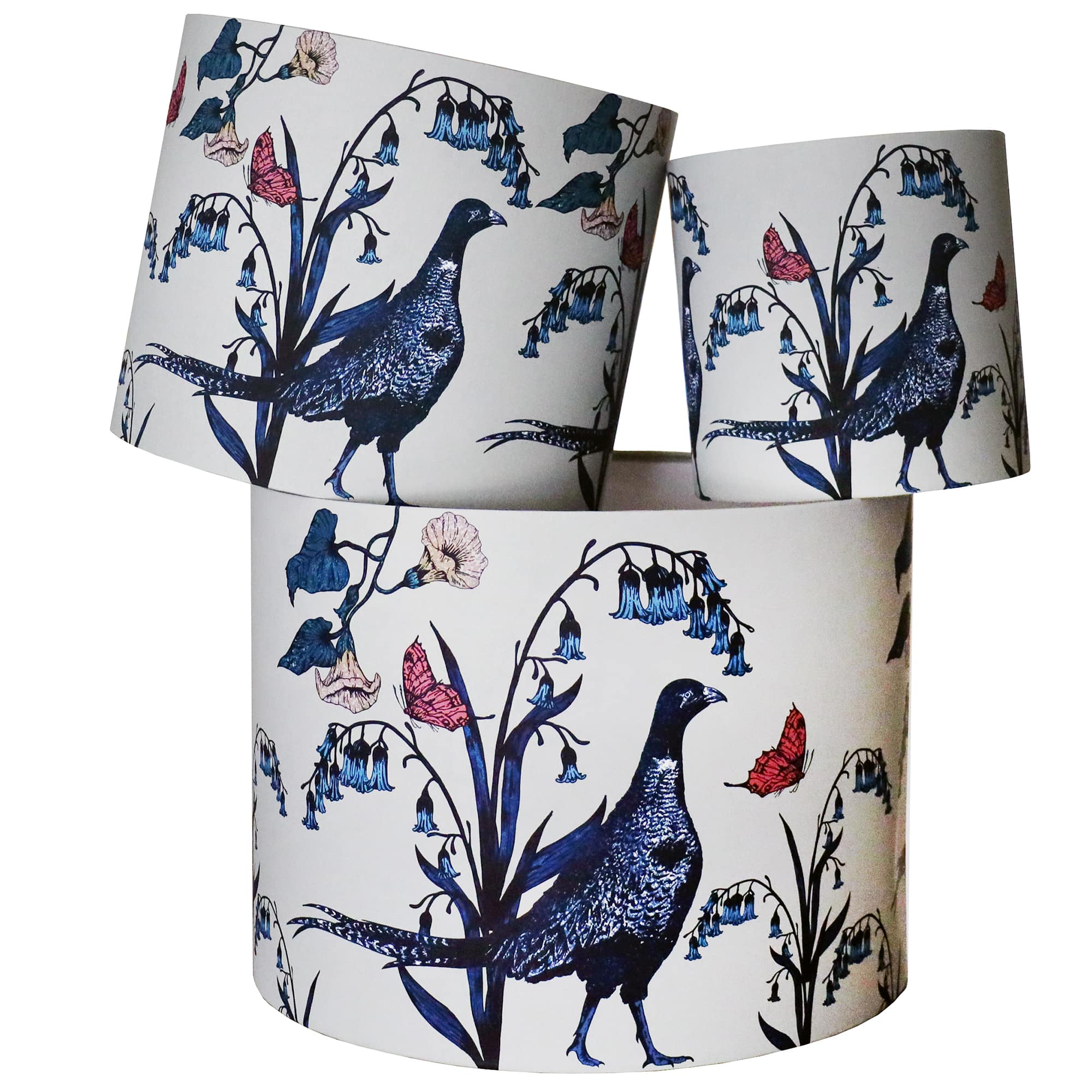 A set of three fabric lampshades featuring an intricate nature-inspired design with pheasants, delicate bluebell flowers, and fluttering butterflies. The illustrations are finely detailed in rich shades of blue, red, and cream, creating a timeless and elegant countryside aesthetic. The lampshades are stacked in a visually striking arrangement, showcasing their unique pattern from different angles.