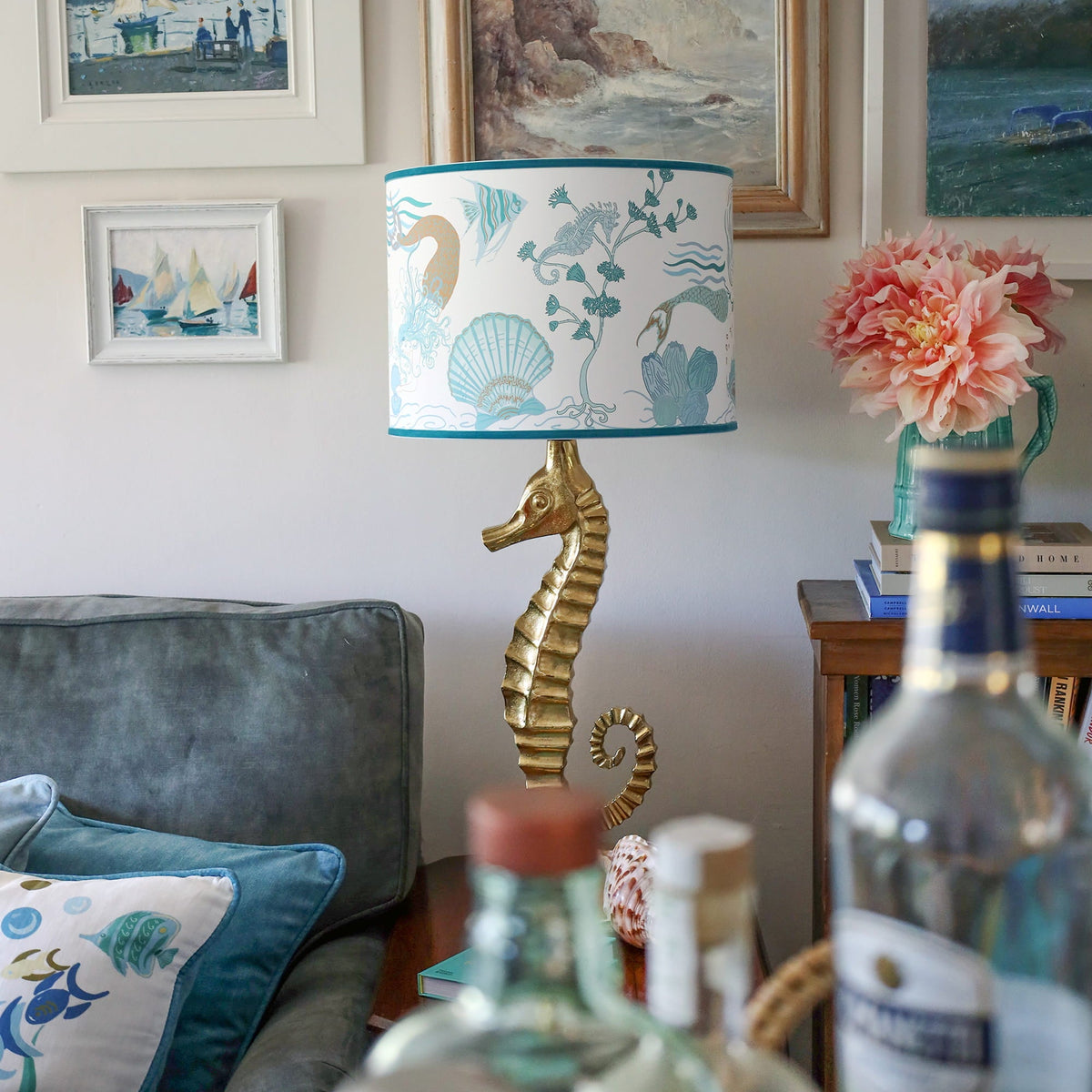 Cream Cornwall Mermaid lampshade on a gold seahorse lamp base, styled with coastal artwork, blue velvet cushions and fresh flowers in a relaxed seaside-inspired living room.