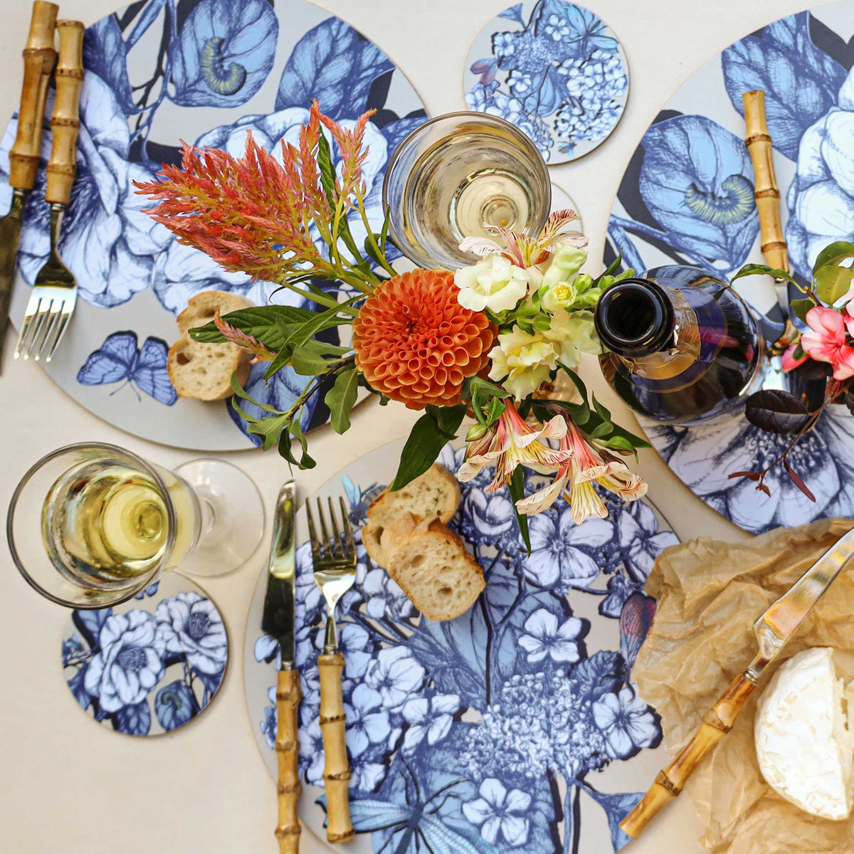 
Overhead view of an elegant table setting featuring round placemats with intricate blue botanical illustrations of flowers, leaves, butterflies and caterpillars on a cream background. A vibrant floral arrangement of orange dahlias, red feathery blooms and delicate cream flowers forms the centrepiece. The setting includes bamboo-handled cutlery, glasses of white wine, a dark wine bottle, slices of crusty bread, soft cheese on brown parchment paper and matching botanical coasters, all arranged on a light lin