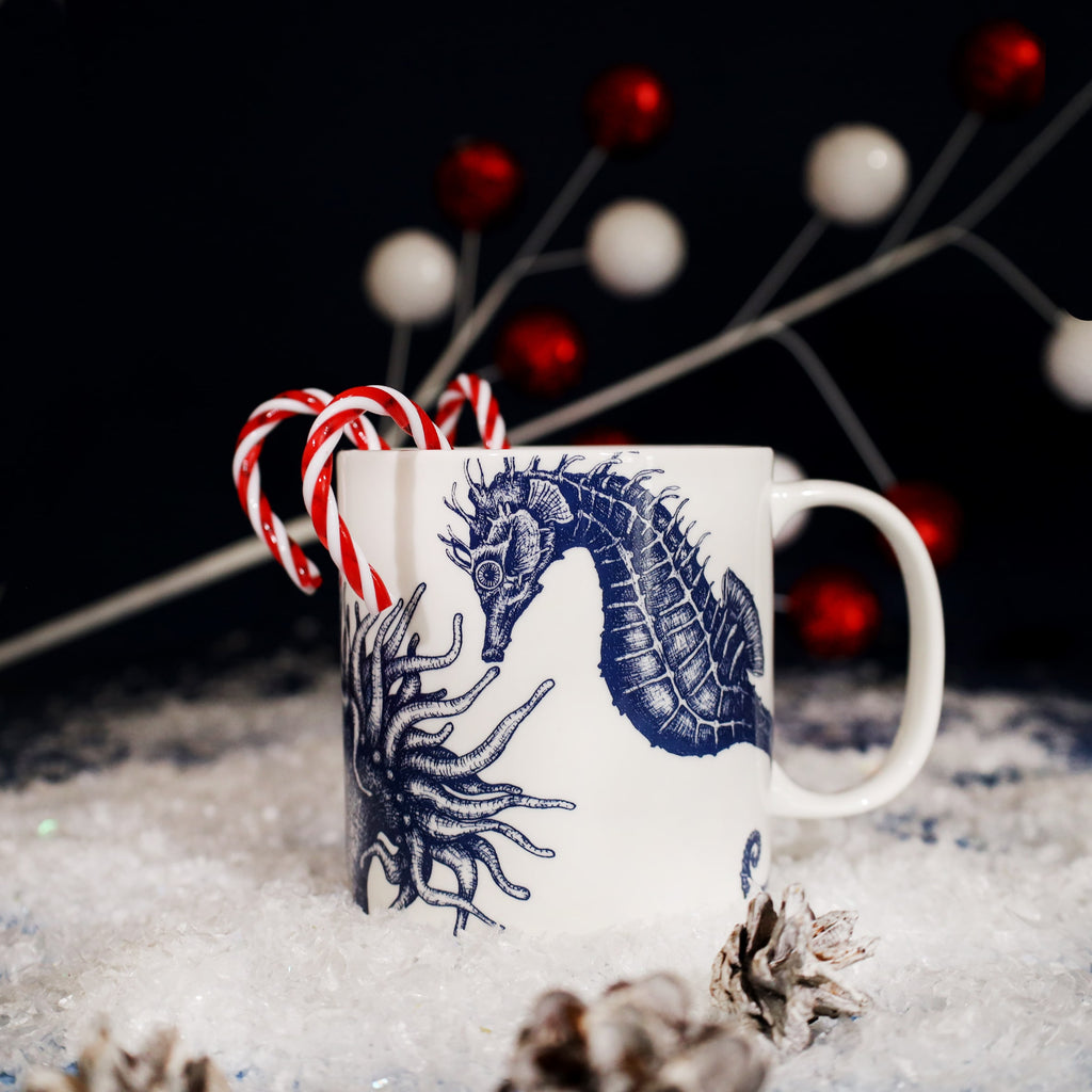 White mug featuring a detailed blue seahorse and anemone illustration, filled with red and white candy canes, set on artificial snow with pinecones and red-and-white festive decorations in the background.