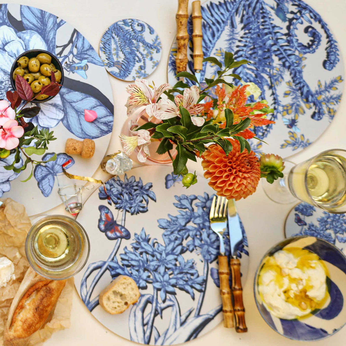Overhead view of an elegant table setting featuring round placemats with intricate blue botanical illustrations of flowers, leaves, ferns, butterflies and insects on a cream background. A vibrant floral centrepiece of orange dahlias, delicate striped cream flowers and bright orange blooms adds colour to the arrangement. Bamboo-handled cutlery, matching botanical coasters, glasses of white wine, a bowl of green olives, crusty bread and soft cheese drizzled with olive oil complete the setting on a light linen