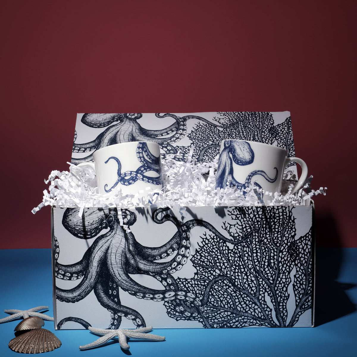 A Cream Cornwall gift set featuring two ceramic mugs with blue octopus designs. The mugs are presented in a matching gift box adorned with intricate octopus and coral illustrations. The box is filled with white shredded paper for cushioning, and decorative seashells and starfish are arranged around it, enhancing the coastal theme against a deep red and blue background