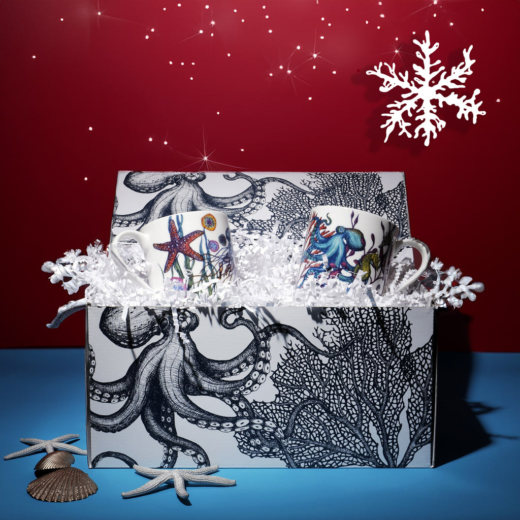 Set of two ocean-themed Reef mugs in a decorative gift box with octopus and coral illustrations, surrounded by white shredded paper and festive snowflake decorations against a red background with twinkling lights.