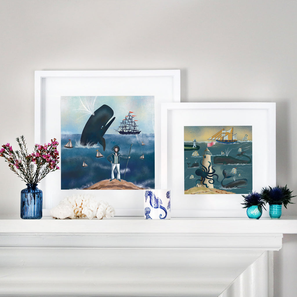 Two framed nautical illustrations displayed on a white mantelpiece. The left artwork features a sailor standing on a rocky shore, gazing at a massive whale leaping from the ocean, surrounded by sailing ships. The right artwork depicts a giant octopus wrapped around a lighthouse, with whales and sailing ships navigating the waves under a golden sky. The mantel is decorated with coral, small blue vases with flowers, and a seahorse-patterned cup.