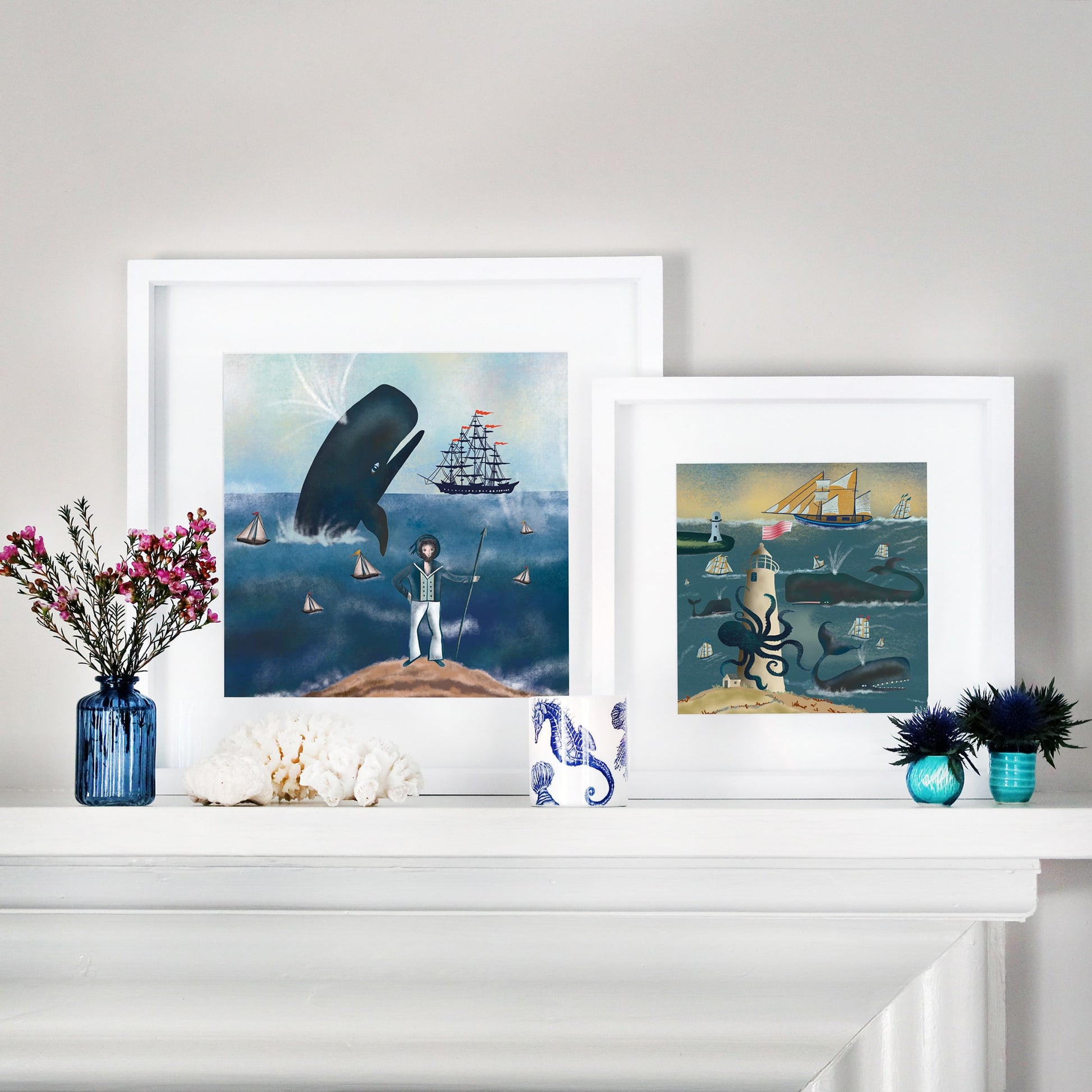 Two framed nautical illustrations displayed on a white mantelpiece. The left artwork features a sailor standing on a rocky shore, gazing at a massive whale leaping from the ocean, surrounded by sailing ships. The right artwork depicts a giant octopus wrapped around a lighthouse, with whales and sailing ships navigating the waves under a golden sky. The mantel is decorated with coral, small blue vases with flowers, and a seahorse-patterned cup.