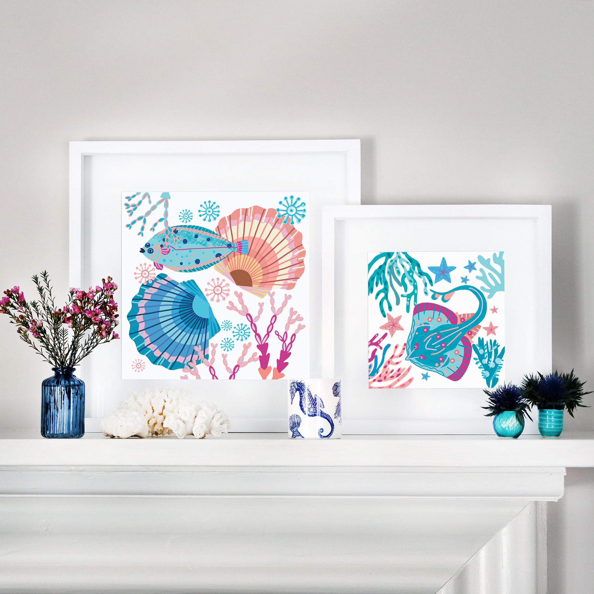  Two framed illustrations with vibrant marine life themes displayed on a white mantelpiece. The left artwork features a colorful flatfish swimming among seashells, coral branches, and sea flora in shades of blue, pink, and orange. The right artwork showcases a stylized stingray or skate surrounded by coral, seaweed, and starfish in a similar bright and whimsical color palette. The frames are complemented by coastal decor, including seashells, flowers, and nautical-themed ceramics.