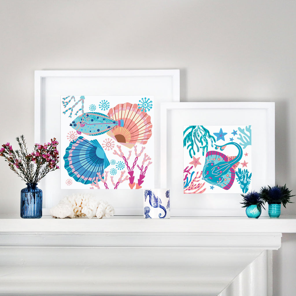 Two framed ocean-themed illustrations displayed on a white mantel. The left frame features a colorful flat fish surrounded by seaweed, coral, and scallop shells in shades of blue, pink, and turquoise. The right frame showcases a vibrant skate swimming among seaweed, starfish, and underwater flora. The scene is accented with coastal decor, including blue glass vases with flowers, a white coral piece, and a ceramic mug with a seahorse design.