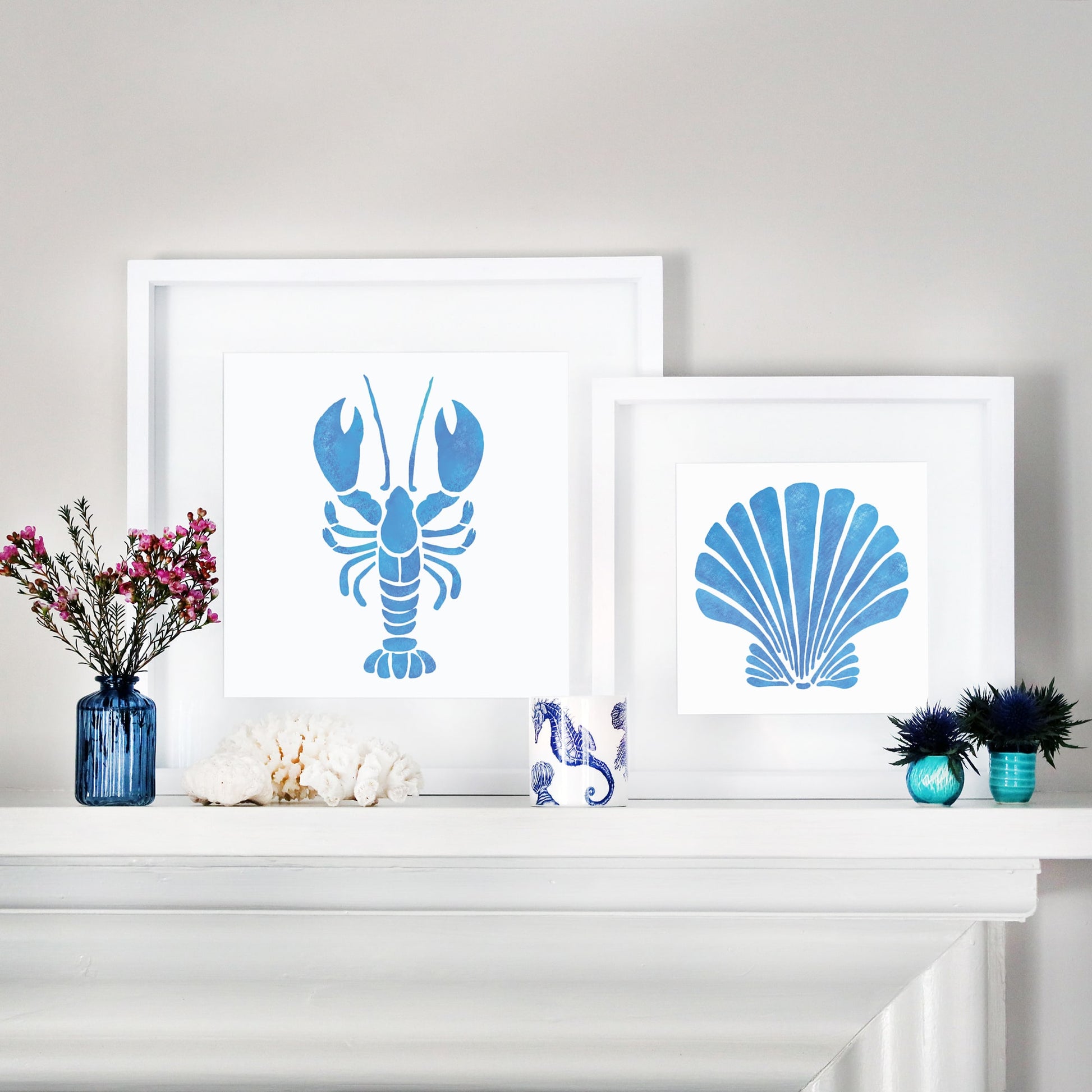 A coastal-inspired mantel display featuring two framed marine-themed artworks. One artwork showcases a bold blue lobster illustration, while the other features a stylized blue scallop shell. The arrangement is complemented by decorative accents, including blue glass vases with flowers, white coral pieces, a seahorse-patterned ceramic cup, and small blue and turquoise vases with greenery. The setup blends oceanic elements with a fresh and modern aesthetic.