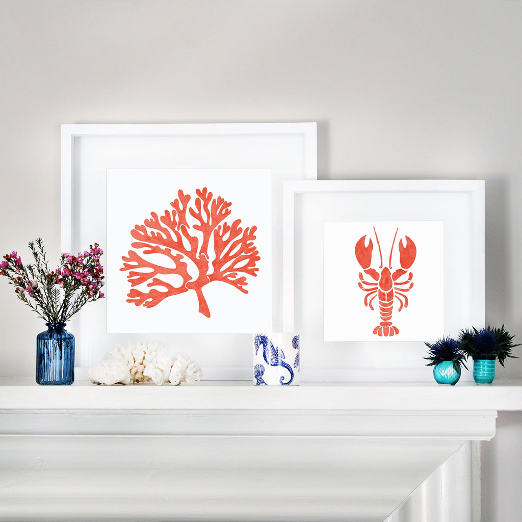 Two framed illustrations, one of an orange coral and the other of an orange lobster, are displayed on a white mantelpiece. The scene is decorated with coastal accents, including a blue vase with pink flowers, coral, seashells, and a blue and white patterned candle holder.
