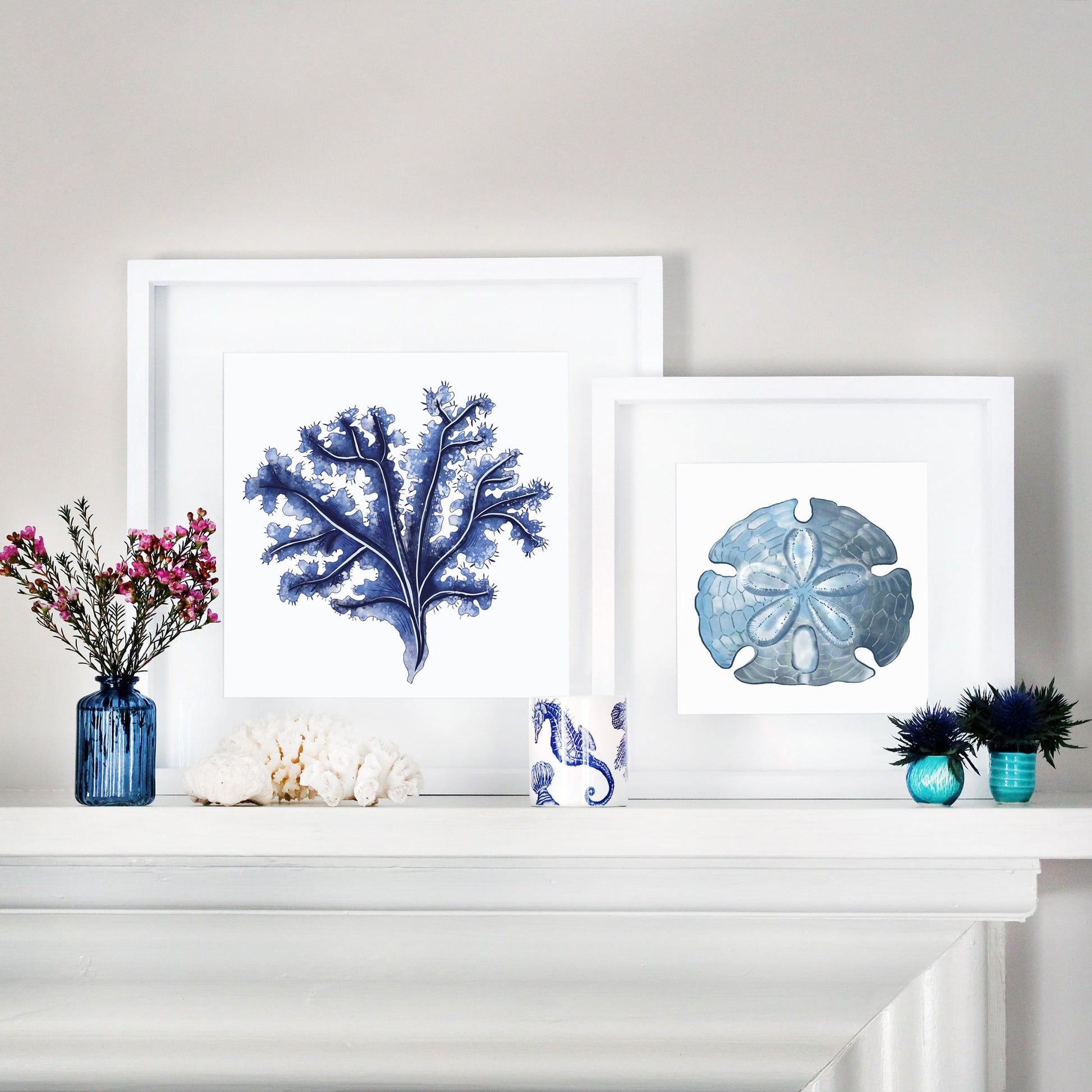 A coastal-inspired mantel display featuring two framed marine-themed artworks. One artwork showcases a detailed blue seaweed illustration, while the other features a blue sand dollar. The arrangement is complemented by decorative accents, including blue glass vases with flowers, white coral pieces, a seahorse-patterned ceramic cup, and small blue and turquoise vases with greenery. The setup blends oceanic elements with a serene and elegant aesthetic.