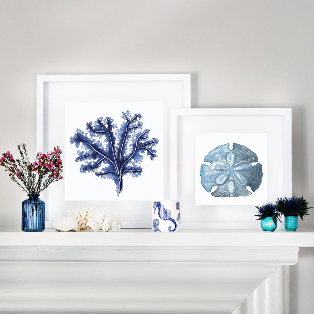 A coastal-inspired mantelpiece display featuring two framed artworks. One frame contains a deep blue seaweed illustration, while the other showcases a delicate sand dollar in shades of blue. The frames are white with a clean, modern mount. The arrangement is complemented by decorative elements, including blue glass vases with flowers, coral pieces, and a candle in a nautical-patterned holder. The scene exudes a serene, oceanic aesthetic.