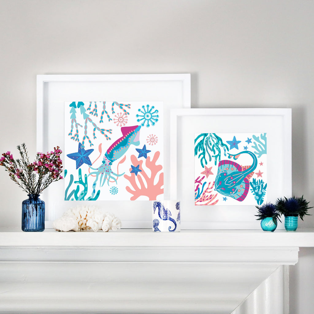 Two framed ocean-themed illustrations displayed on a white mantel. The left frame features a colorful squid swimming among coral, starfish, and seaweed in shades of blue, pink, and turquoise. The right frame showcases a vibrant skate surrounded by underwater flora and marine life. The mantel is decorated with a blue glass vase with pink flowers, white coral pieces, a ceramic cup with a seahorse design, and small blue and turquoise floral arrangements.