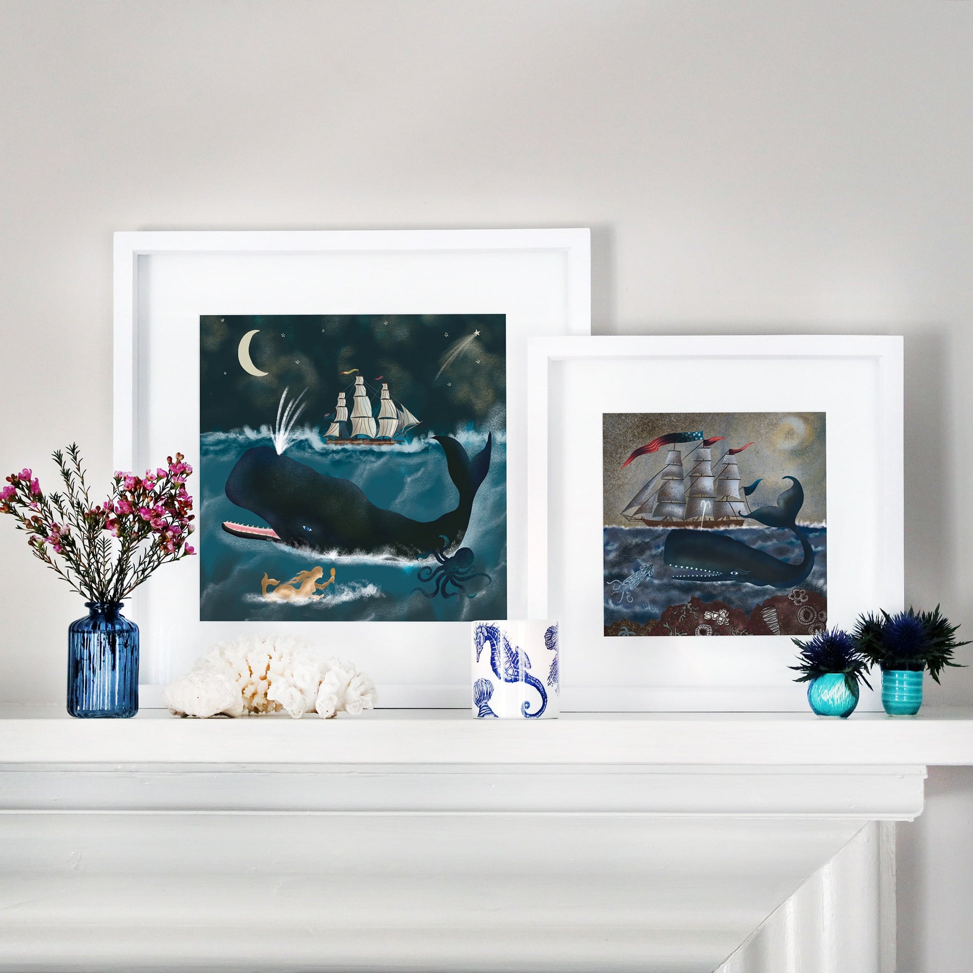 Two framed ocean-themed illustrations displayed on a white mantel. The left frame features a large whale, a sailing ship under a crescent moon, a mermaid, and an octopus beneath the waves. The right frame depicts a whale with a ship on the horizon, surrounded by sea creatures and barnacles. The mantel is decorated with blue glass vases, coral, and a candle in a nautical-themed holder.