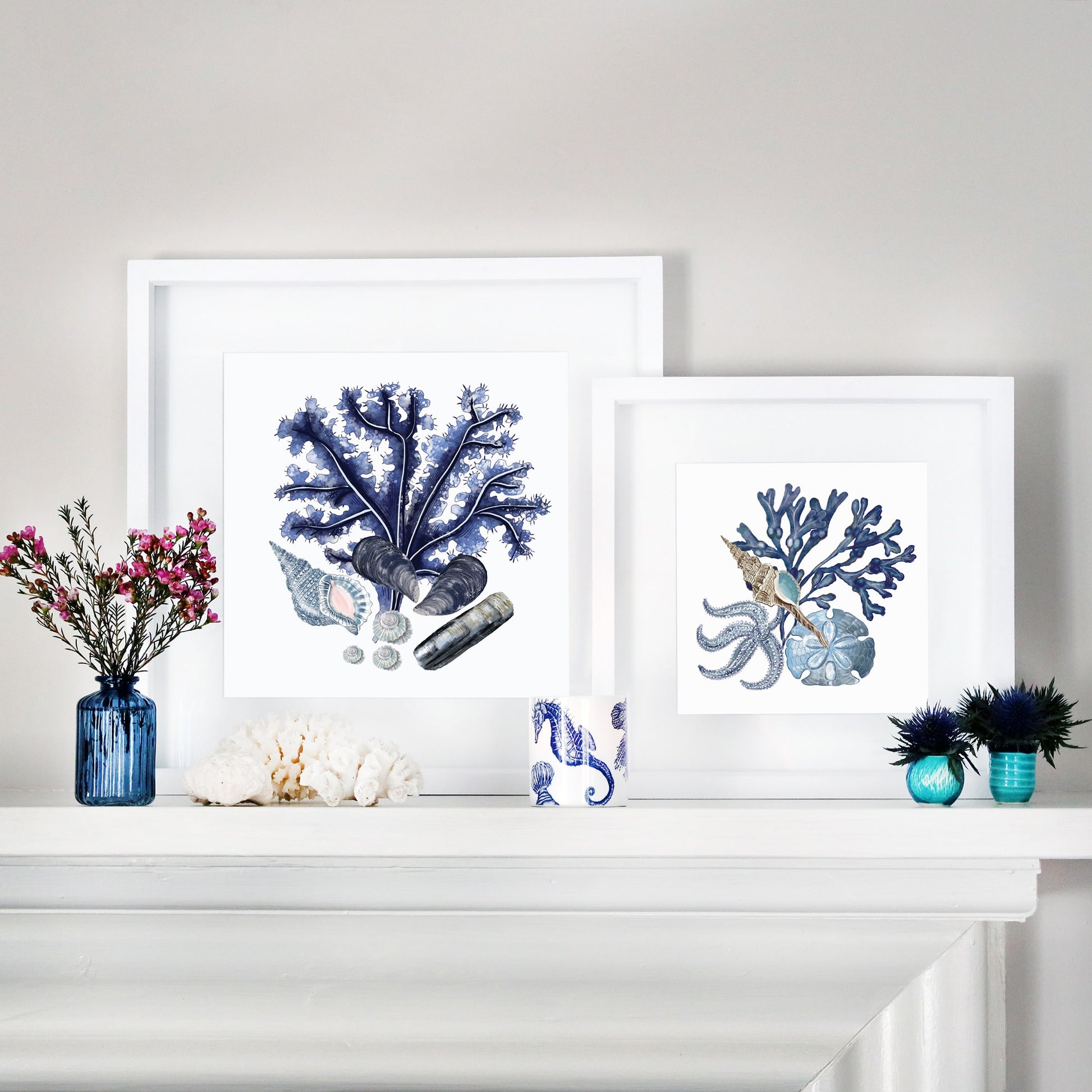 A coastal-themed mantel display featuring two framed marine-inspired artworks with blue coral, seashells, and starfish illustrations. The frames are layered for depth, with decorative accents including small blue glass vases with flowers, a white coral piece, and a ceramic cup with a seahorse design. The setup combines soft blues and neutral tones for a serene, ocean-inspired aesthetic.