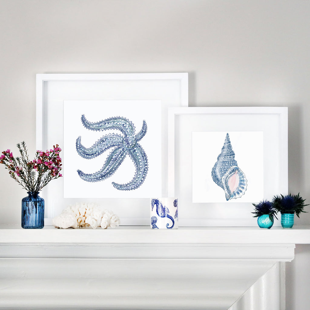 A coastal-inspired mantel display featuring two framed marine-themed artworks. One artwork showcases a detailed blue starfish, while the other features a blue whelk shell with intricate textures and soft pink accents. The arrangement is complemented by decorative accents, including blue glass vases with flowers, white coral pieces, a seahorse-patterned ceramic cup, and small blue and turquoise vases with greenery. The setup blends oceanic elements with a serene and elegant aesthetic.