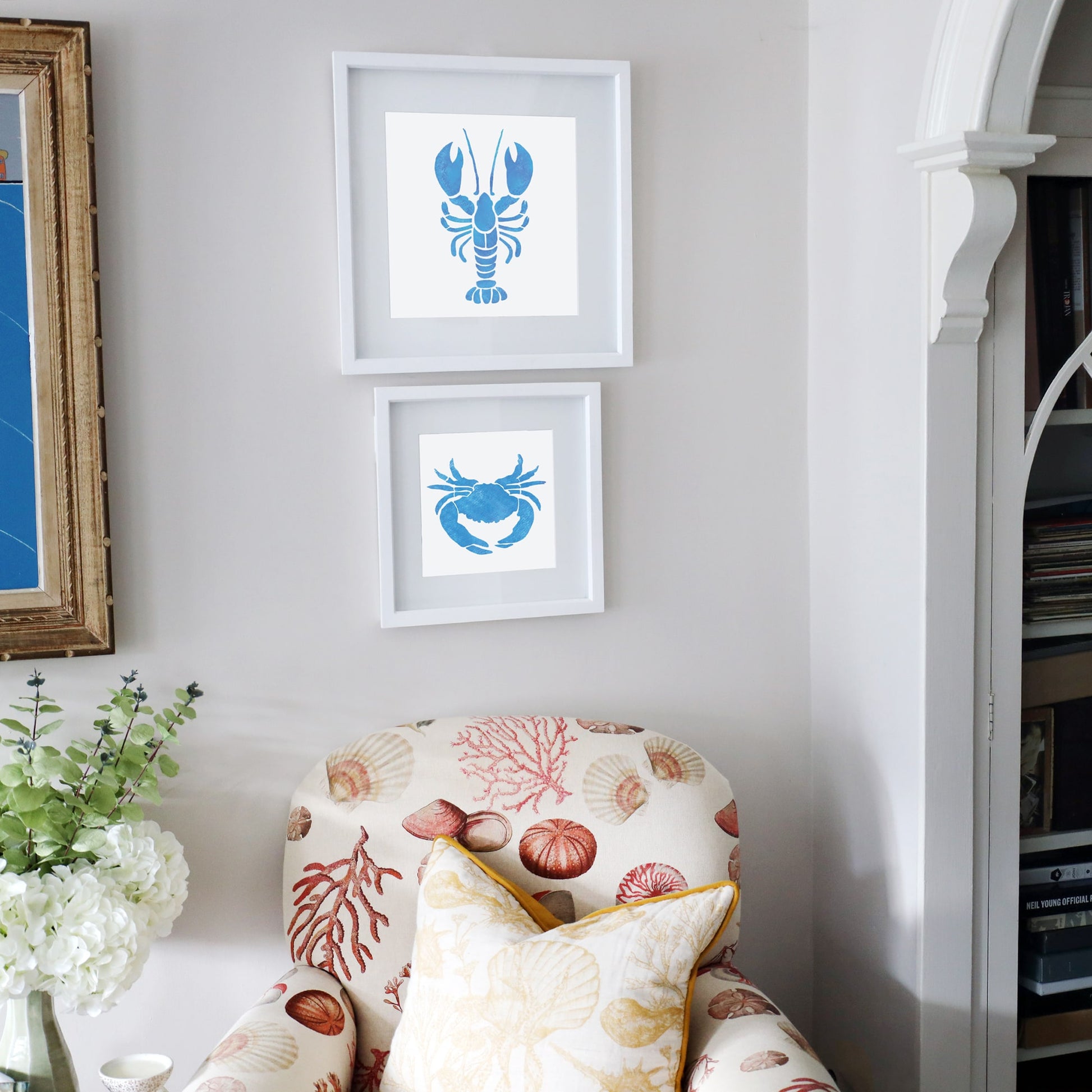 A cozy reading nook with a coastal theme, featuring a seashell-patterned armchair in warm coral and cream tones, complemented by a yellow-trimmed cushion. Above the chair, two framed marine-inspired artworks hang on the wall—one depicting a bold blue lobster silhouette and the other showcasing a stylized blue crab. A side table with a vase of white hydrangeas and greenery enhances the calming, ocean-inspired atmosphere.
