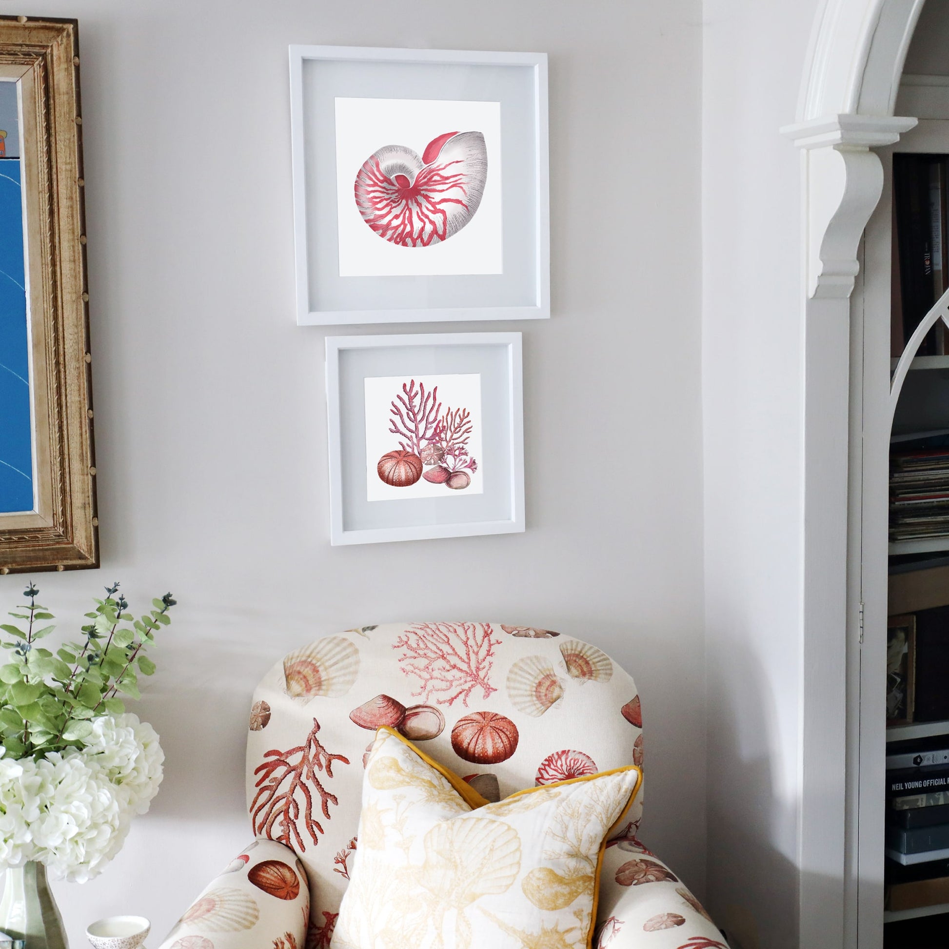 A cozy reading nook featuring a seashell-patterned armchair in warm coral and cream tones, paired with a yellow-trimmed cushion. Above, two framed coastal-themed artworks are arranged on the wall—one depicting a detailed nautilus shell and the other featuring red coral, a sea urchin, a sand dollar, and seashells. A side table with a vase of white hydrangeas and greenery adds a fresh touch to the serene, ocean-inspired space.