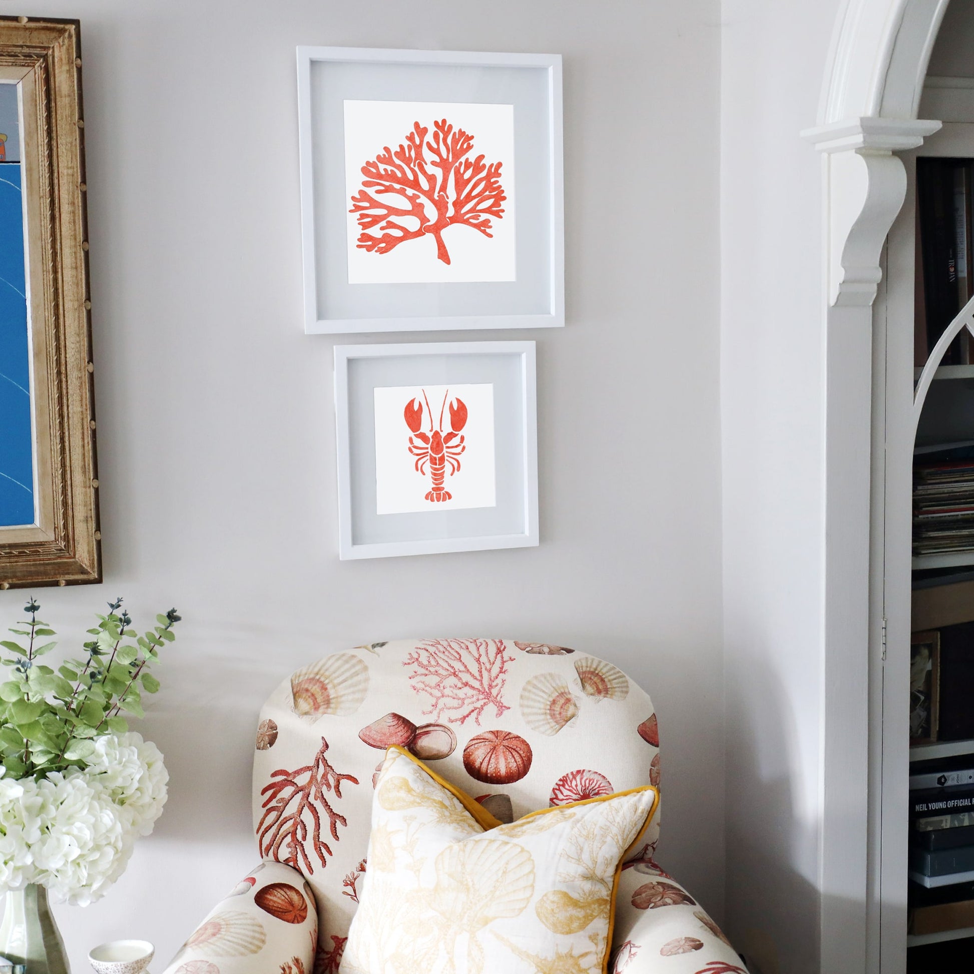 A cozy coastal-themed corner featuring a patterned armchair with seashell and coral motifs, a cream and yellow cushion, and two framed artworks on the wall. The top frame showcases an orange coral illustration, while the bottom frame displays an orange lobster silhouette. The neutral-toned wall and soft furnishings create a relaxed, beach-inspired aesthetic.