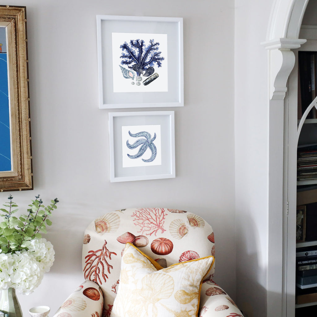 Two framed coastal prints featuring blue coral and a starfish, hung above a seashell-patterned armchair with a yellow-trimmed cushion in a light-filled, nautical-inspired living space.