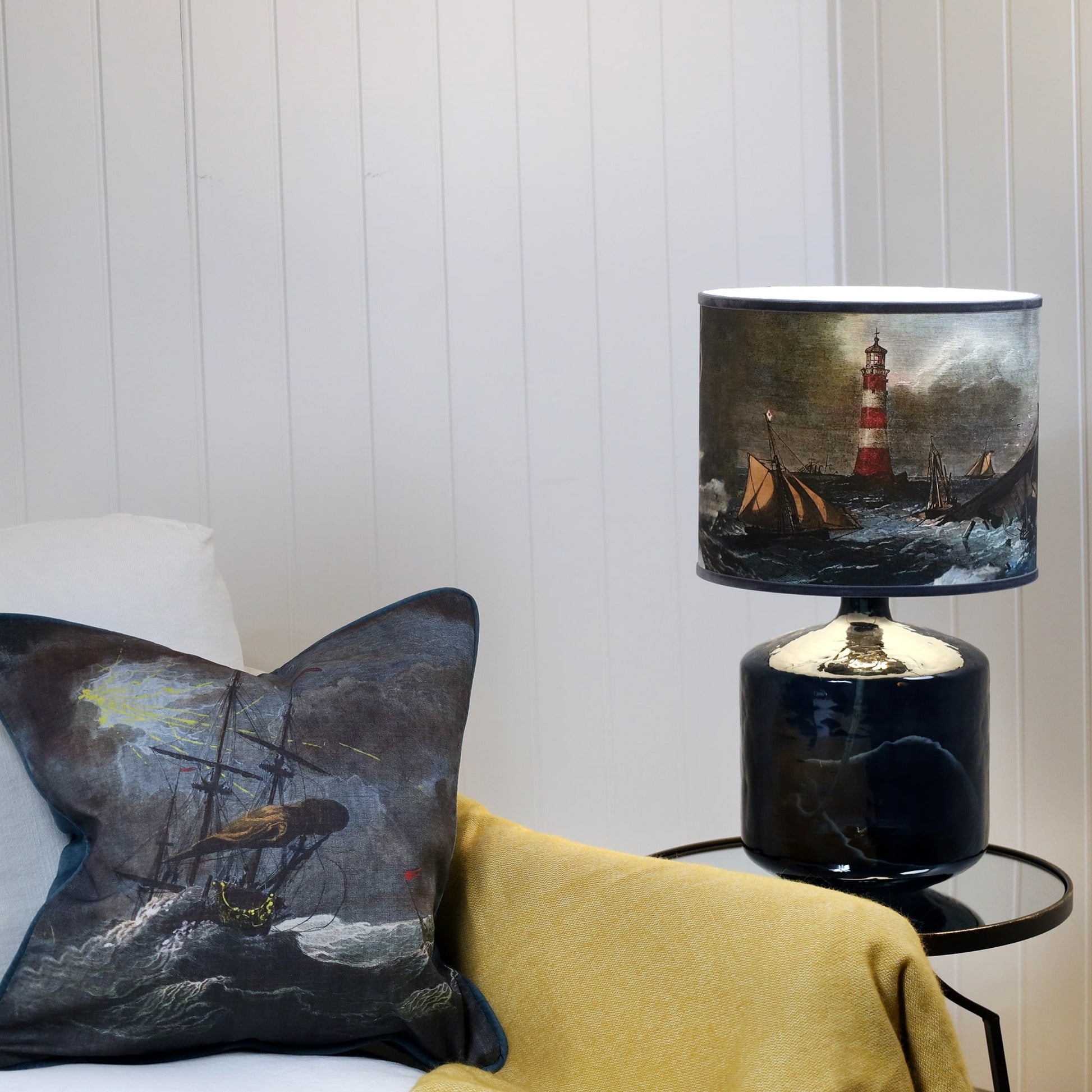A shipwreck scene lampshade in blues & brown sitting on a petrol blue glass lamp base, set against a white shiplap wall a mustard throw over the chair and the corner of a matching cushion.