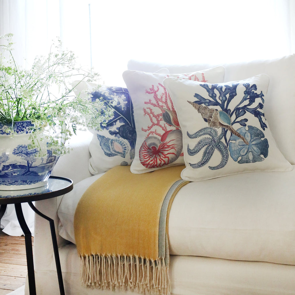 3 cushions an a white sofa with starfish, shells, seaweeds and sand dollar illustration in tones of blues & pinks on with the sun streaming through the window at the back and a large willow pattern jug filled with cow parsley