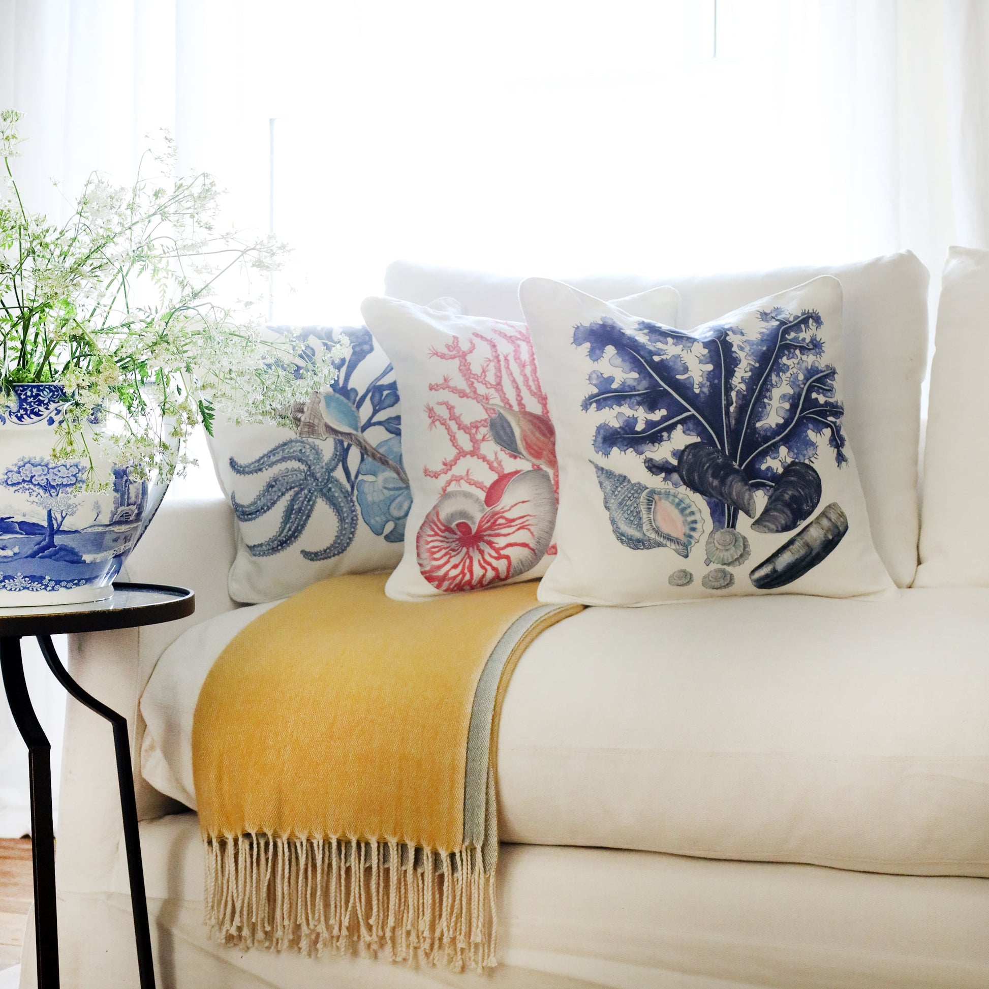 3 cushions an a white sofa with starfish, shells, seaweeds and sand dollar illustration in tones of blues & pinks on with the sun streaming through the window at the back and a large willow pattern jug filled with cow parsley