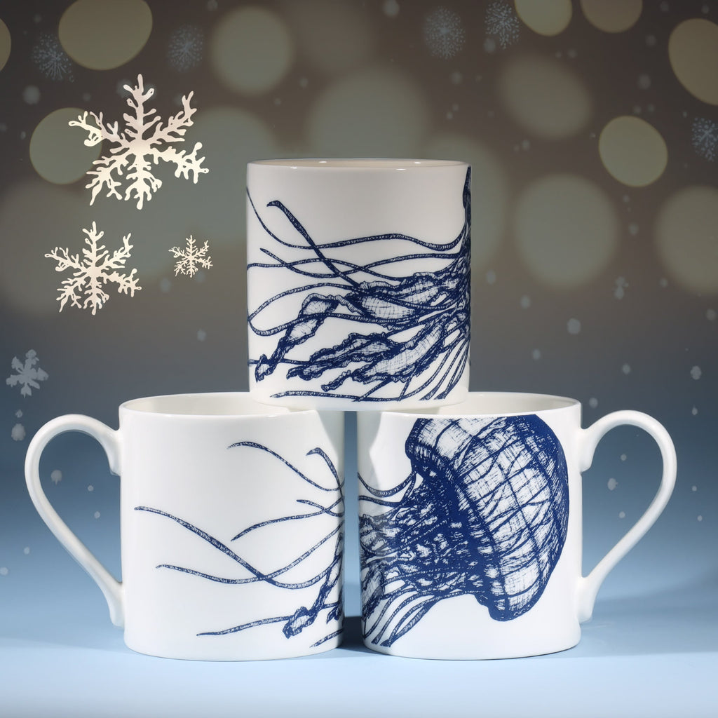 Three white mugs featuring intricate blue jellyfish illustrations arranged in a pyramid shape, set against a softly lit festive background with delicate snowflakes and small, warm golden lights.