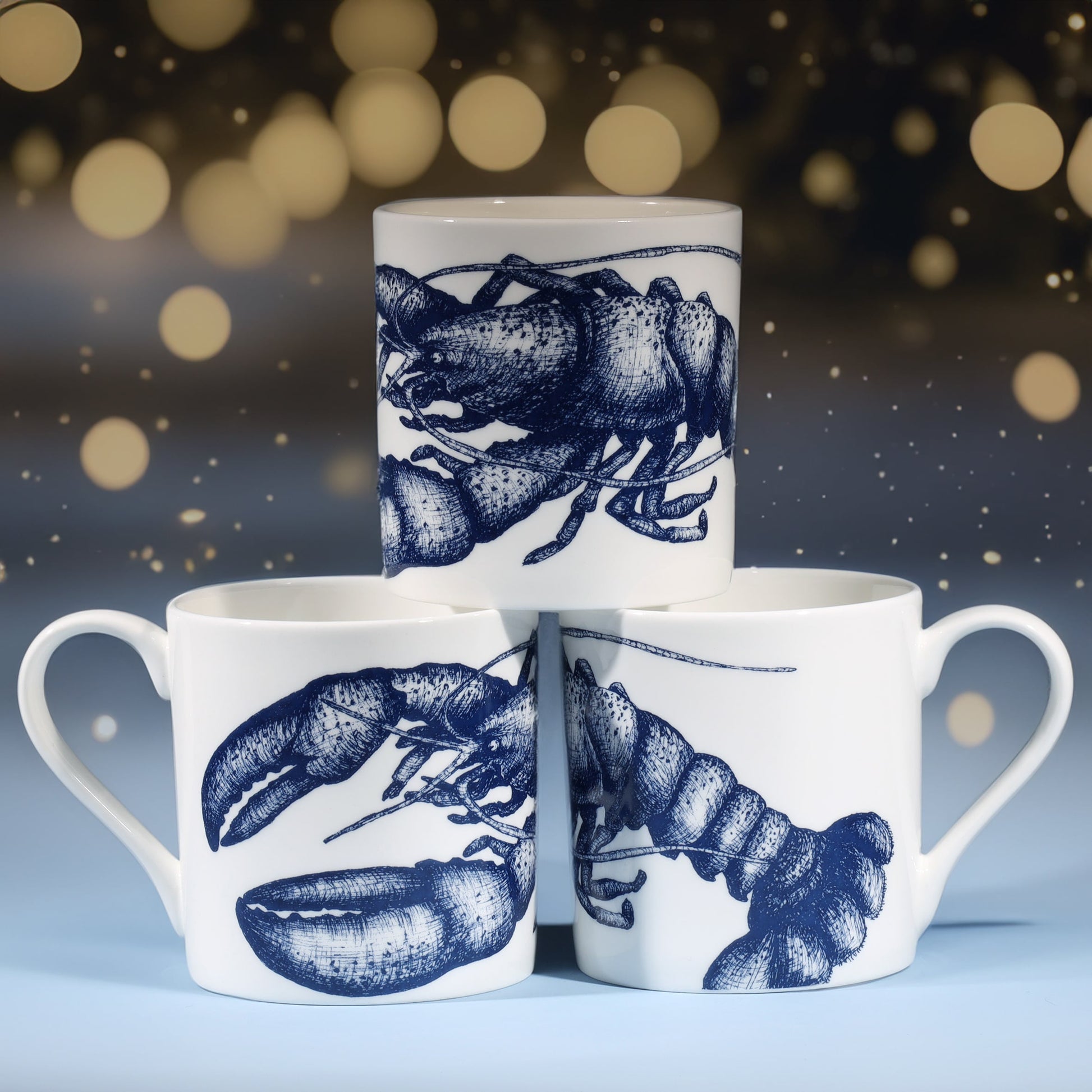 Three white mugs featuring intricate navy blue lobster illustrations arranged in a pyramid stack, set against a softly lit background with warm, golden twinkling lights and a cool blue base for a festive winter feel.