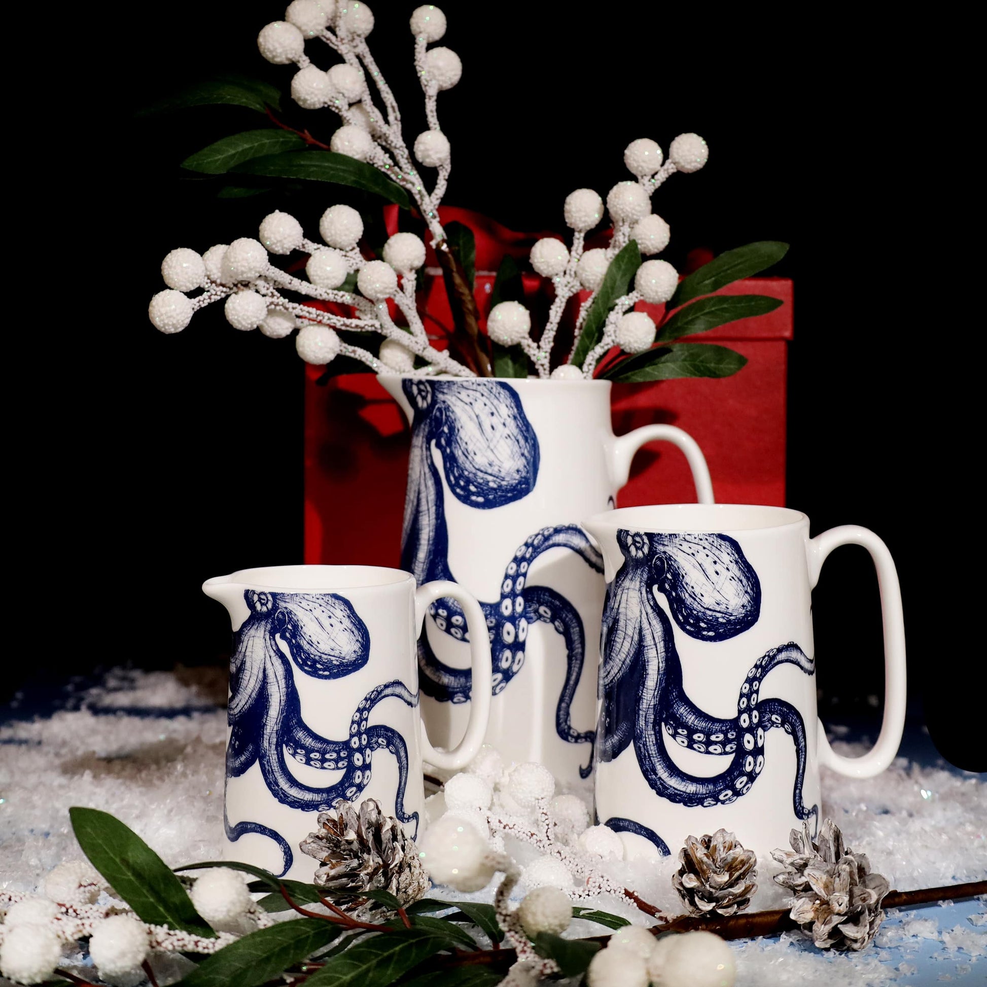 Three white ceramic jugs featuring intricate blue octopus illustrations, arranged on artificial snow with pinecones, green foliage, and white berry decorations, set against a black background with a red gift box for a festive display.