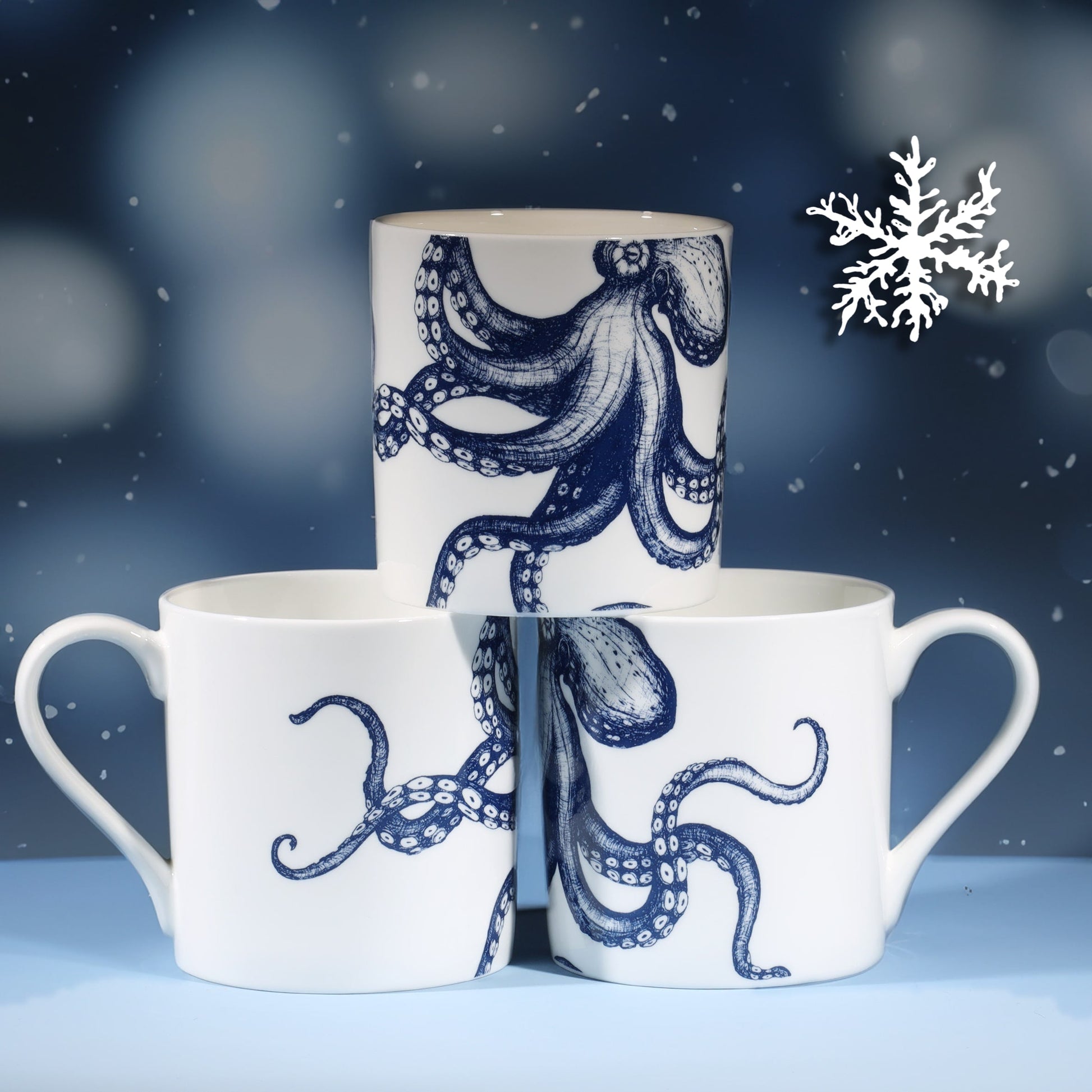 Three white ceramic mugs featuring a navy octopus illustration are stacked in a pyramid against a cool blue background with softly blurred lights and falling snowflakes, creating a festive winter feel.
