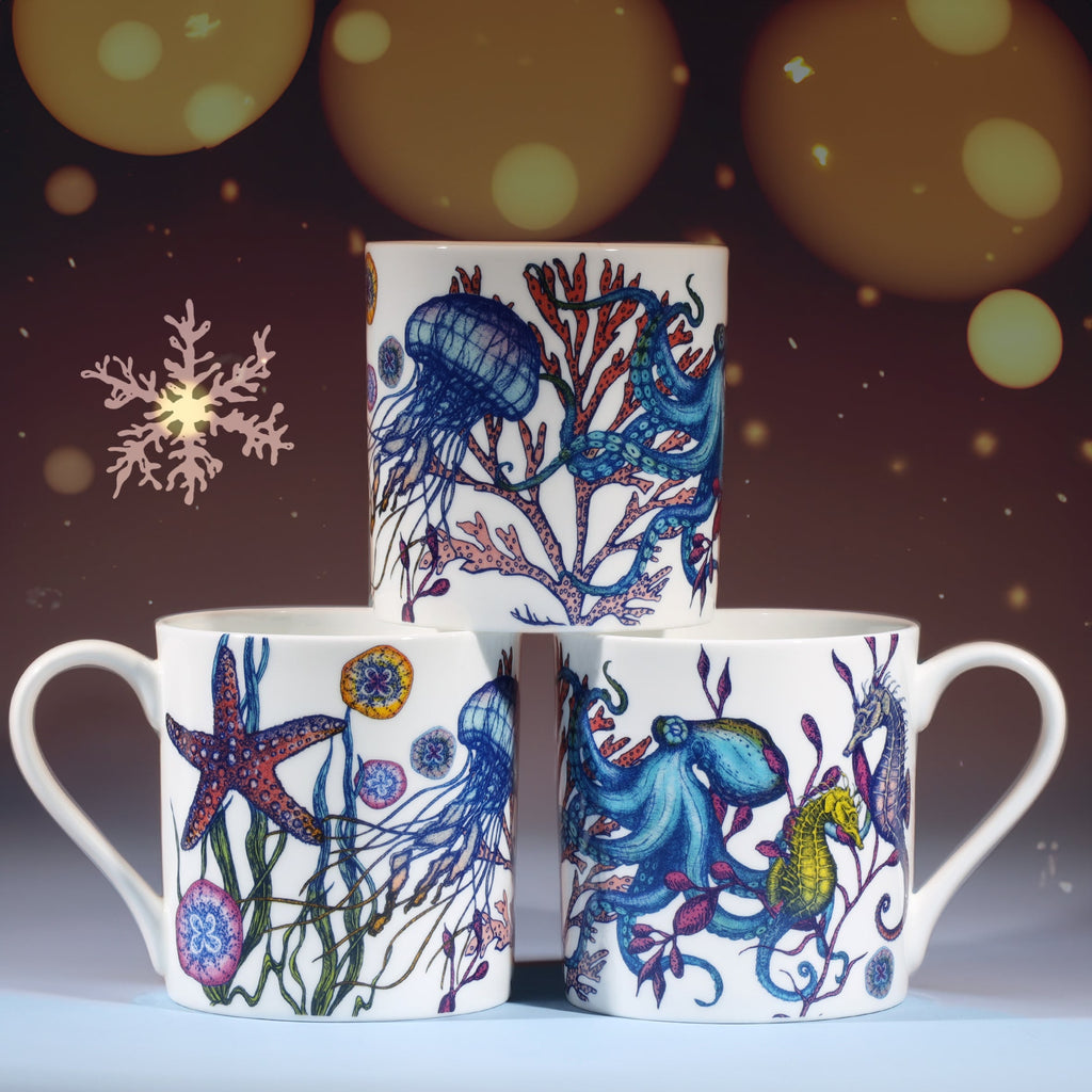 Three white mugs decorated with colorful marine illustrations of octopus, jellyfish, seahorse, and starfish, stacked in a pyramid against a warm festive background with softly glowing golden lights and a delicate snowflake design.