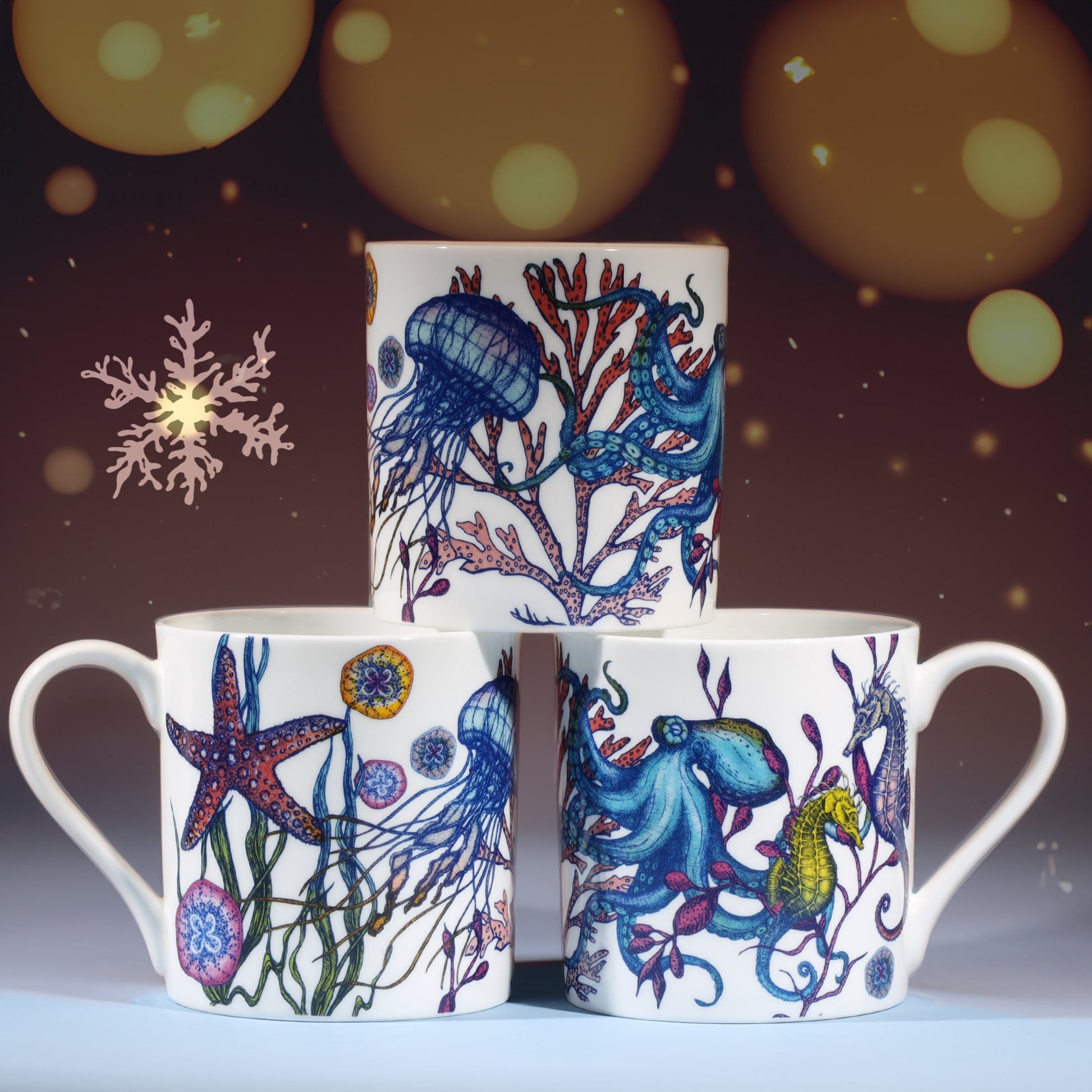Three white mugs decorated with colorful marine illustrations of octopus, jellyfish, seahorse, and starfish, stacked in a pyramid against a warm festive background with softly glowing golden lights and a delicate snowflake design.