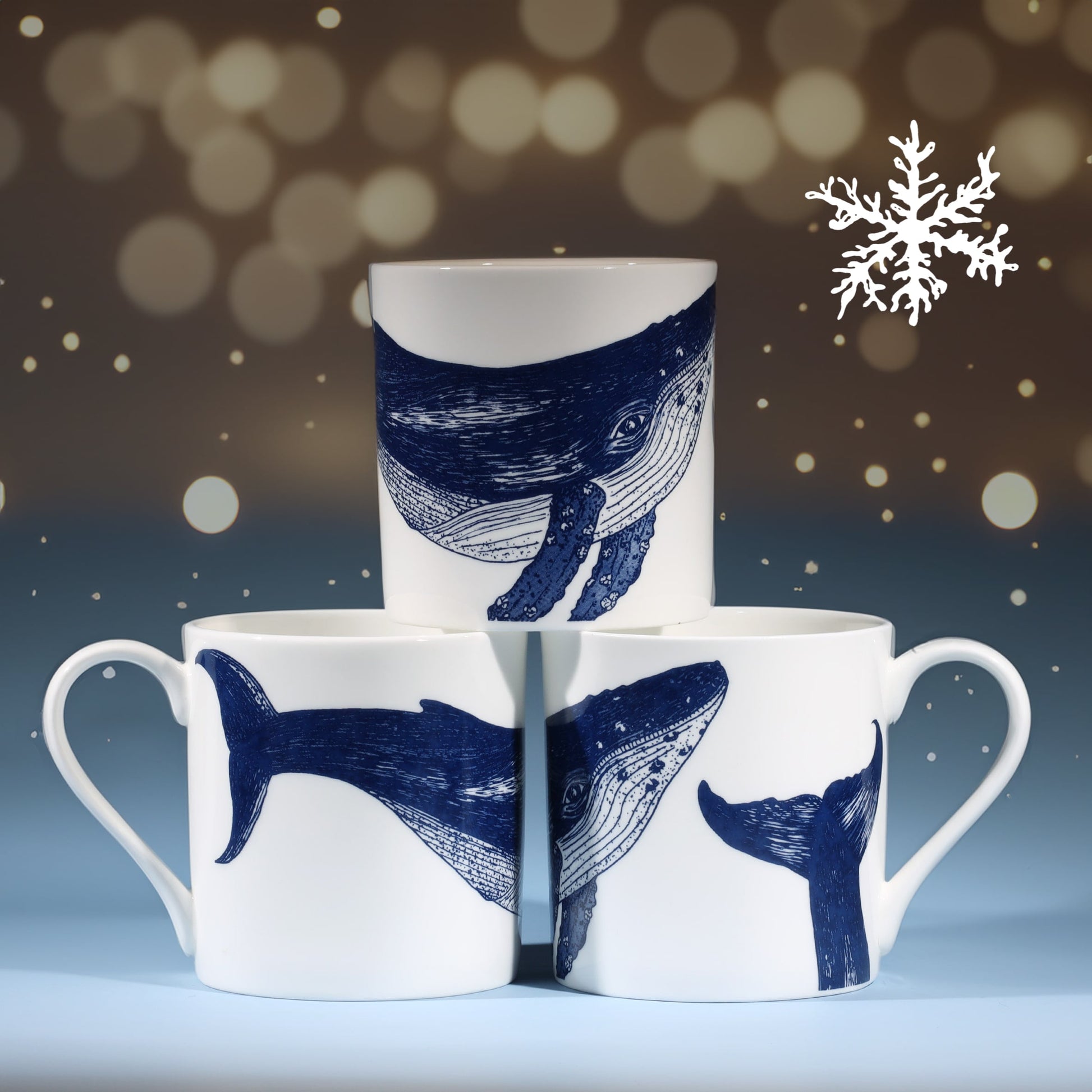 Three white mugs decorated with detailed navy blue whale illustrations arranged in a pyramid formation, with a softly glowing background of small golden lights and a white snowflake, creating a festive winter atmosphere.