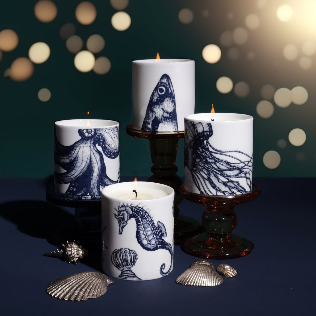 Set of four lit candles in white holders decorated with navy blue sea life illustrations, including a seahorse, octopus, fish, and jellyfish, displayed with seashells on a dark blue surface against a softly glowing festive background.