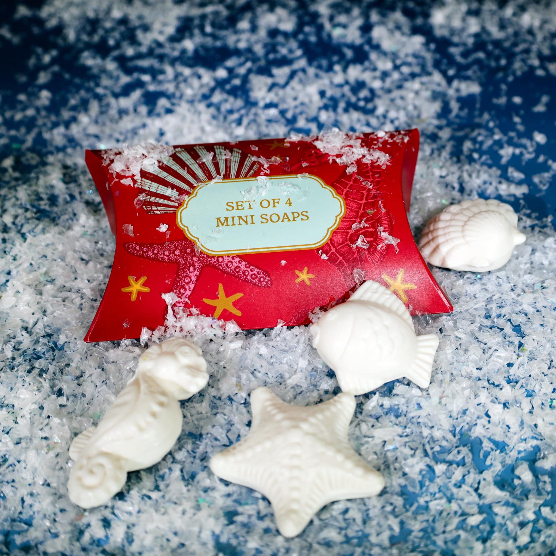 Festive red box labeled "Set of 4 Mini Soaps" surrounded by four white soaps shaped like a seahorse, starfish, fish, and seashell, displayed on a bed of artificial snow with a wintry blue background.