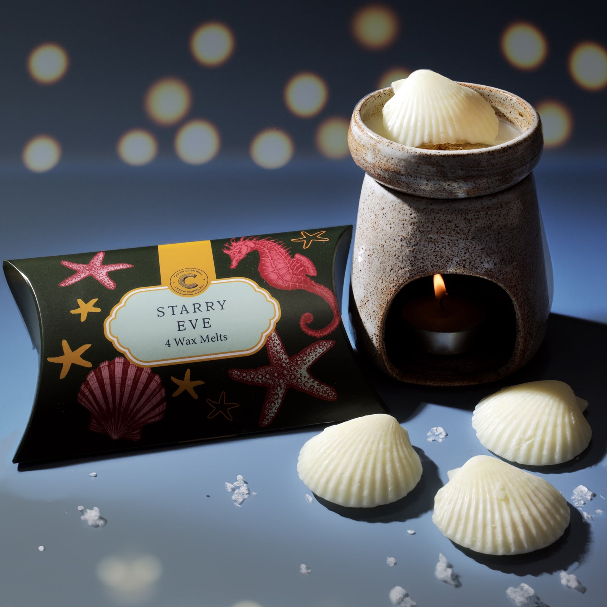 Cream Cornwall Starry Eve wax melts displayed with a ceramic wax burner; the dark green box decorated with red sea creatures sits beside shell-shaped white wax melts, one melting atop the burner with a tealight glowing beneath, set against a softly lit blue background.