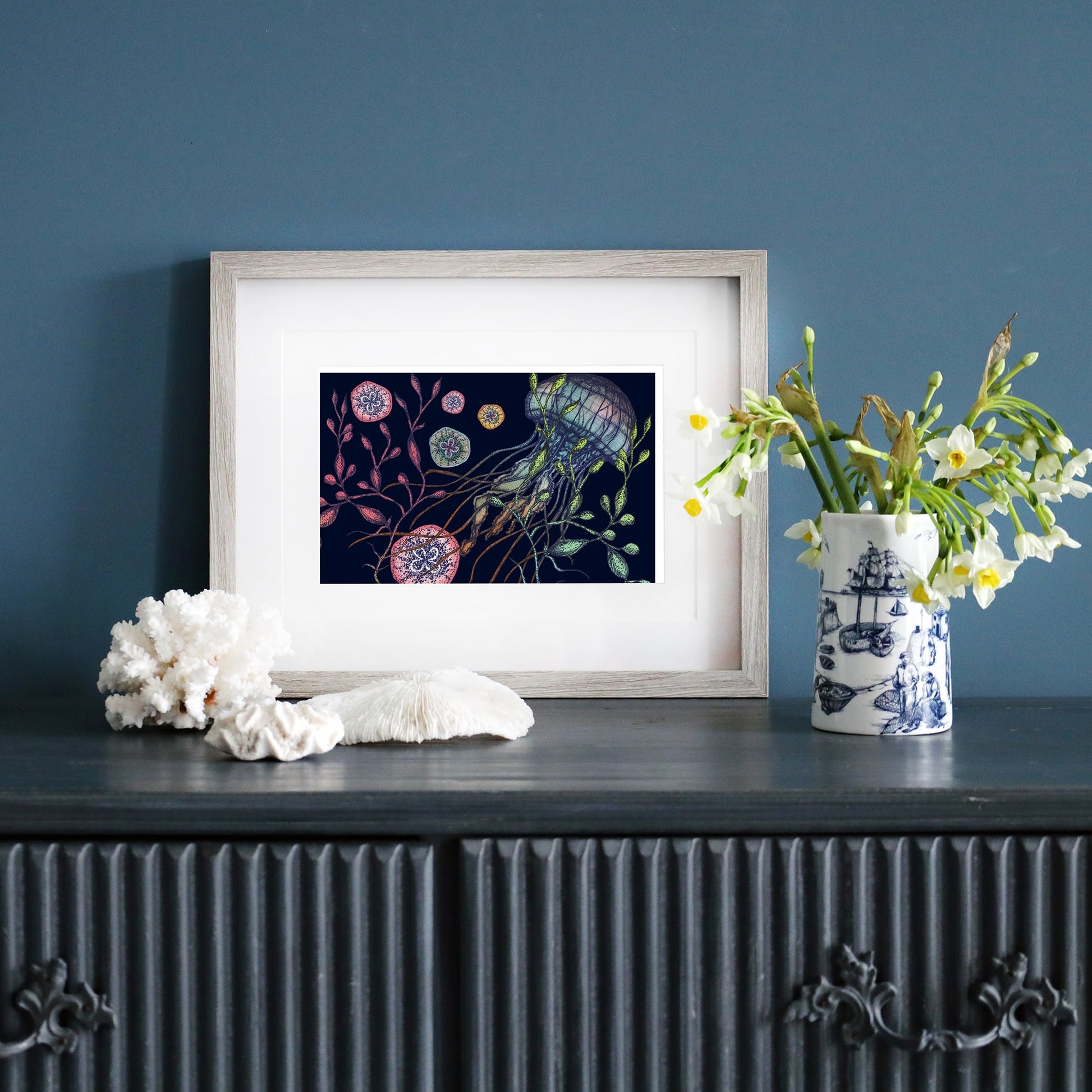 A framed illustration of a jellyfish with seaweed and marine elements on a dark navy background, set in a pale wood frame with a white mount. The artwork is displayed on a dark sideboard, accompanied by coral, a vase of white flowers, and a blue and white nautical-themed ceramic jug.