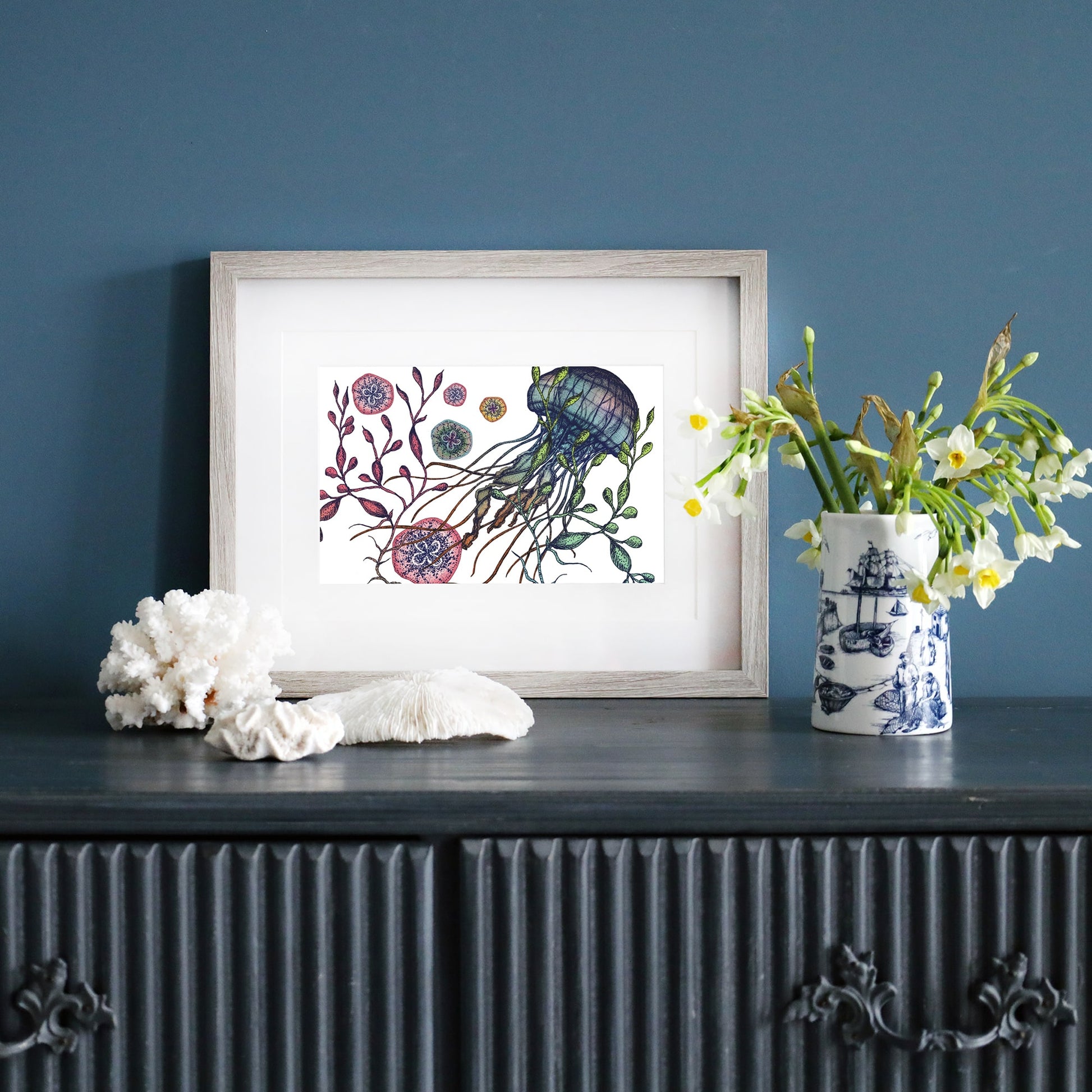 A framed jellyfish illustration in a light wood frame sits on a dark sideboard against a deep blue wall. Next to it, a vase with nautical designs holds white flowers, while coral pieces are arranged nearby.