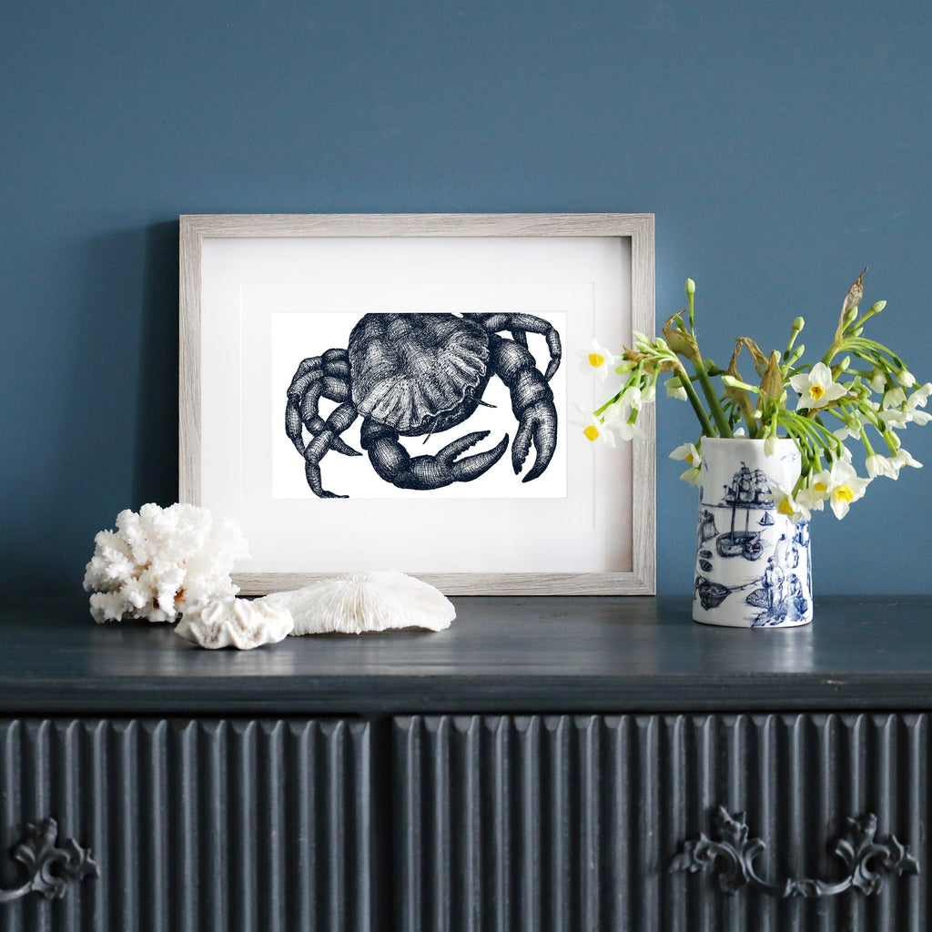 A framed blue-and-white illustration of a crab in a pale wood frame, displayed on a dark blue wall. Below, a dark sideboard is decorated with white coral, a ceramic vase with yellow and white flowers, and a blue and white nautical-patterned mug.