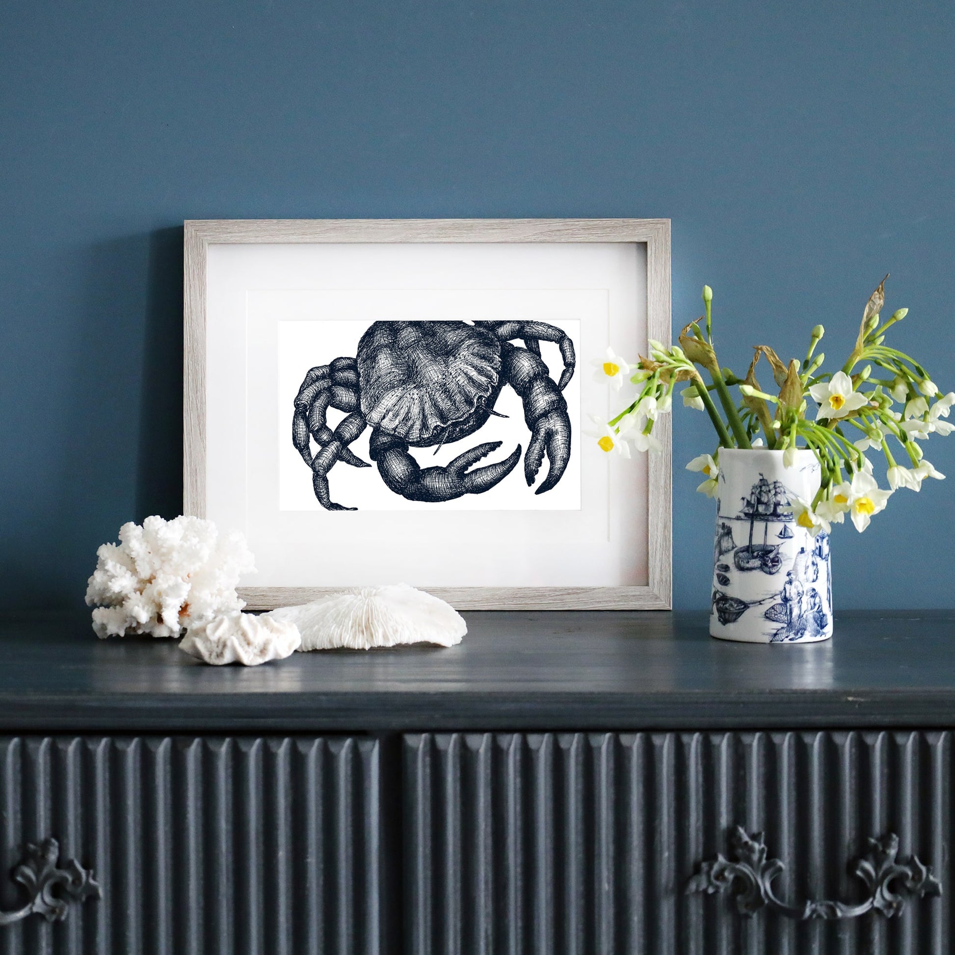 A framed blue-and-white illustration of a crab in a pale wood frame, displayed on a dark blue wall. Below, a dark sideboard is decorated with white coral, a ceramic vase with yellow and white flowers, and a blue and white nautical-patterned mug.