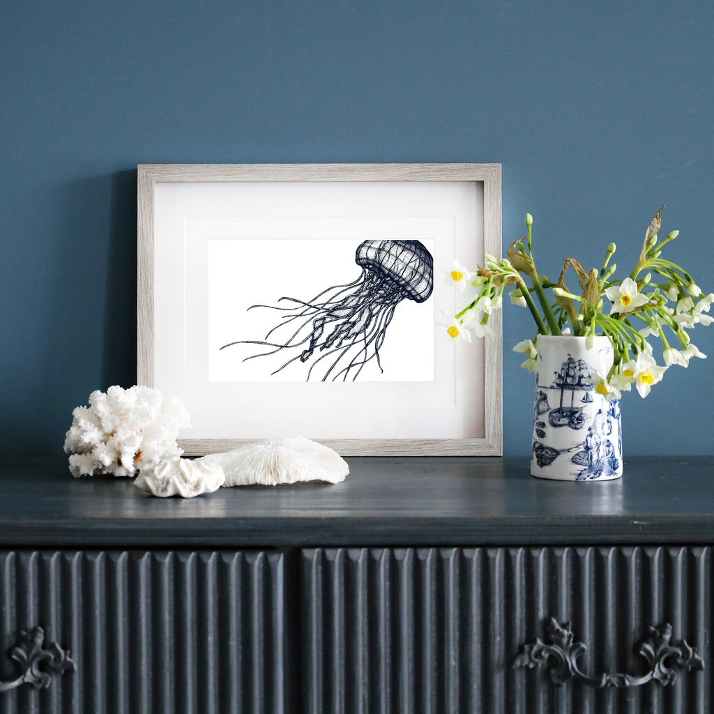 A framed blue-and-white illustration of a jellyfish in a pale wood frame, displayed on a dark blue wall. Below, a dark sideboard is decorated with white coral, a ceramic vase with yellow and white flowers, and a blue and white nautical-patterned mug.