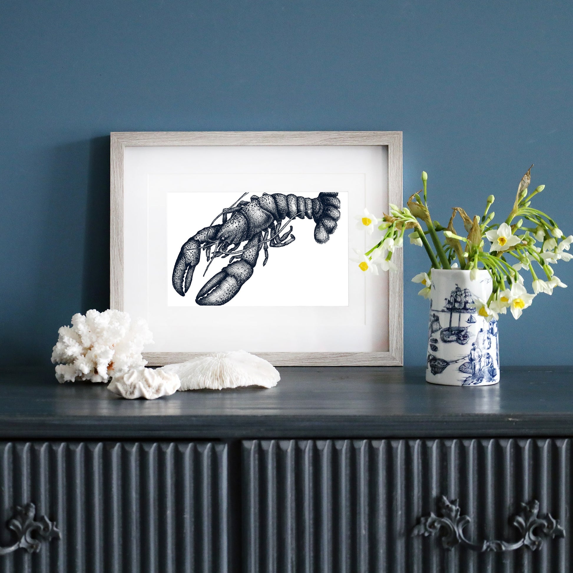 A framed blue-and-white illustration of a lobster in a pale wood frame, displayed on a dark blue wall. Below, a dark sideboard is decorated with white coral, a ceramic vase with yellow and white flowers, and a blue and white nautical-patterned mug.