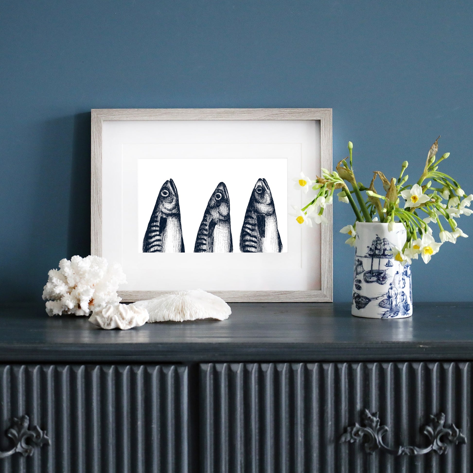 A framed blue-and-white illustration of 3 mackerel heads in a pale wood frame, displayed on a dark blue wall. Below, a dark sideboard is decorated with white coral, a ceramic vase with yellow and white flowers, and a blue and white nautical-patterned mug.