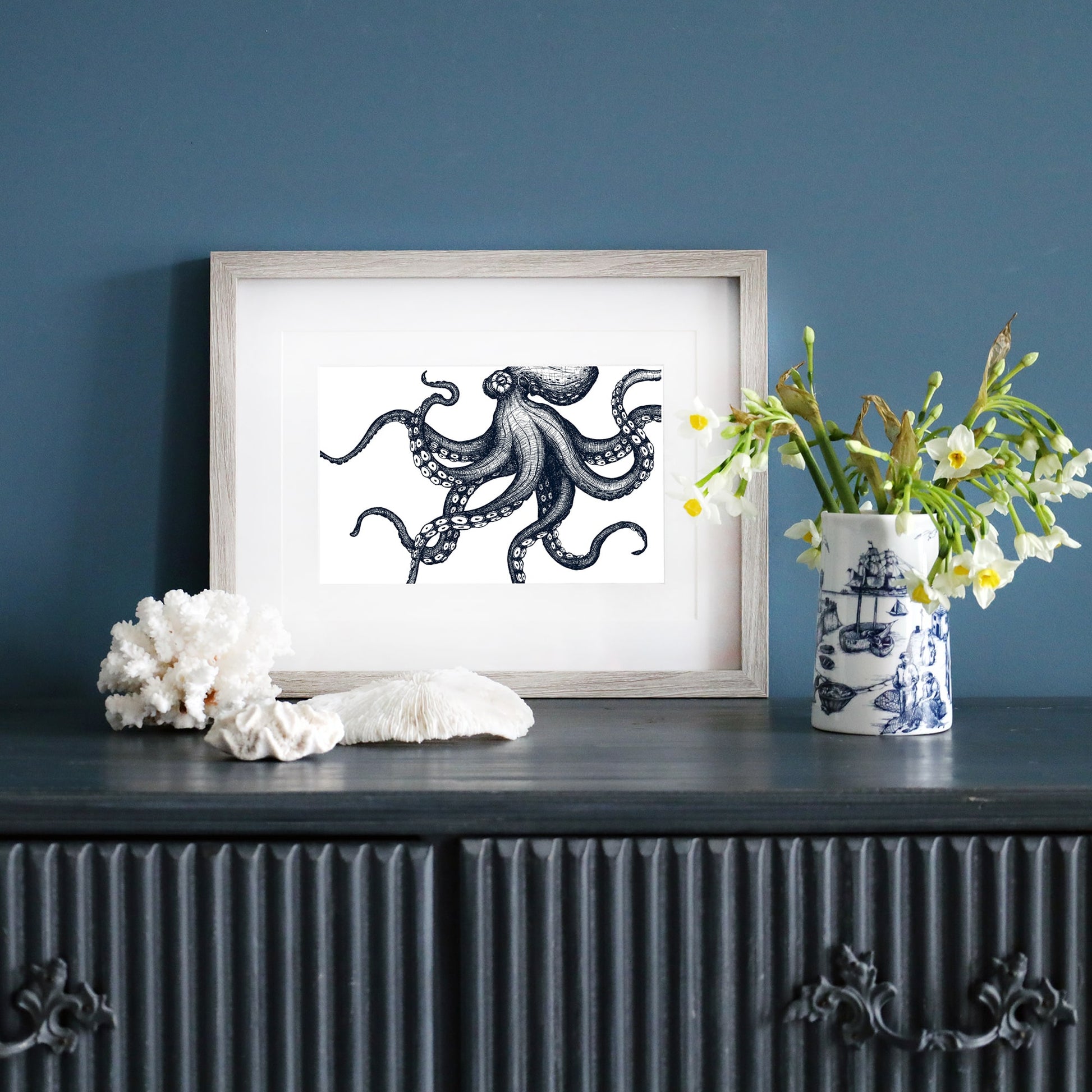 A framed black-and-white illustration of an octopus in a pale wood frame, displayed on a dark blue wall. Below, a dark sideboard is decorated with white coral, a ceramic vase with yellow and white flowers, and a blue and white nautical-patterned mug.