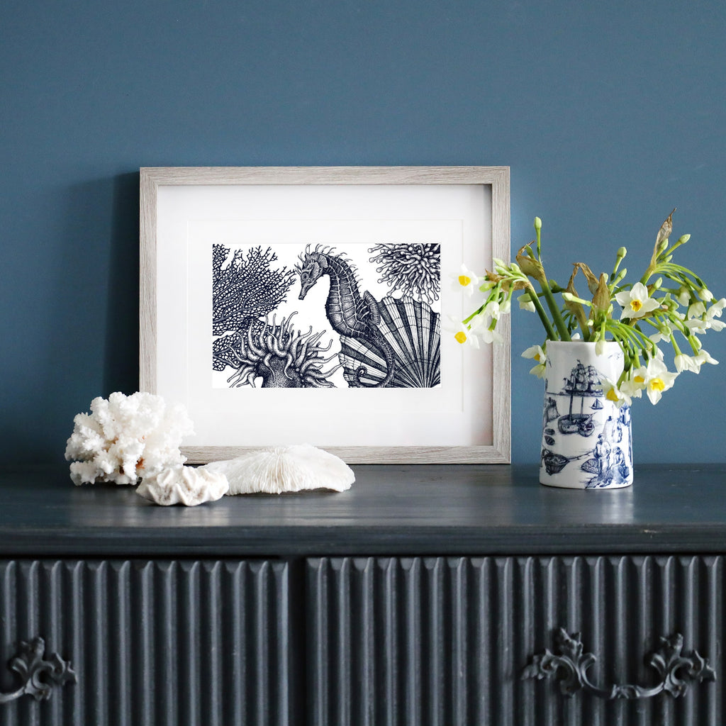 A framed black-and-white illustration of an underwater scene featuring a seahorse, coral, and sea anemones, displayed on a dark wood sideboard against a deep blue wall. The sideboard is decorated with white coral pieces, a ceramic vase with fresh flowers, and a blue-and-white maritime-themed mug.