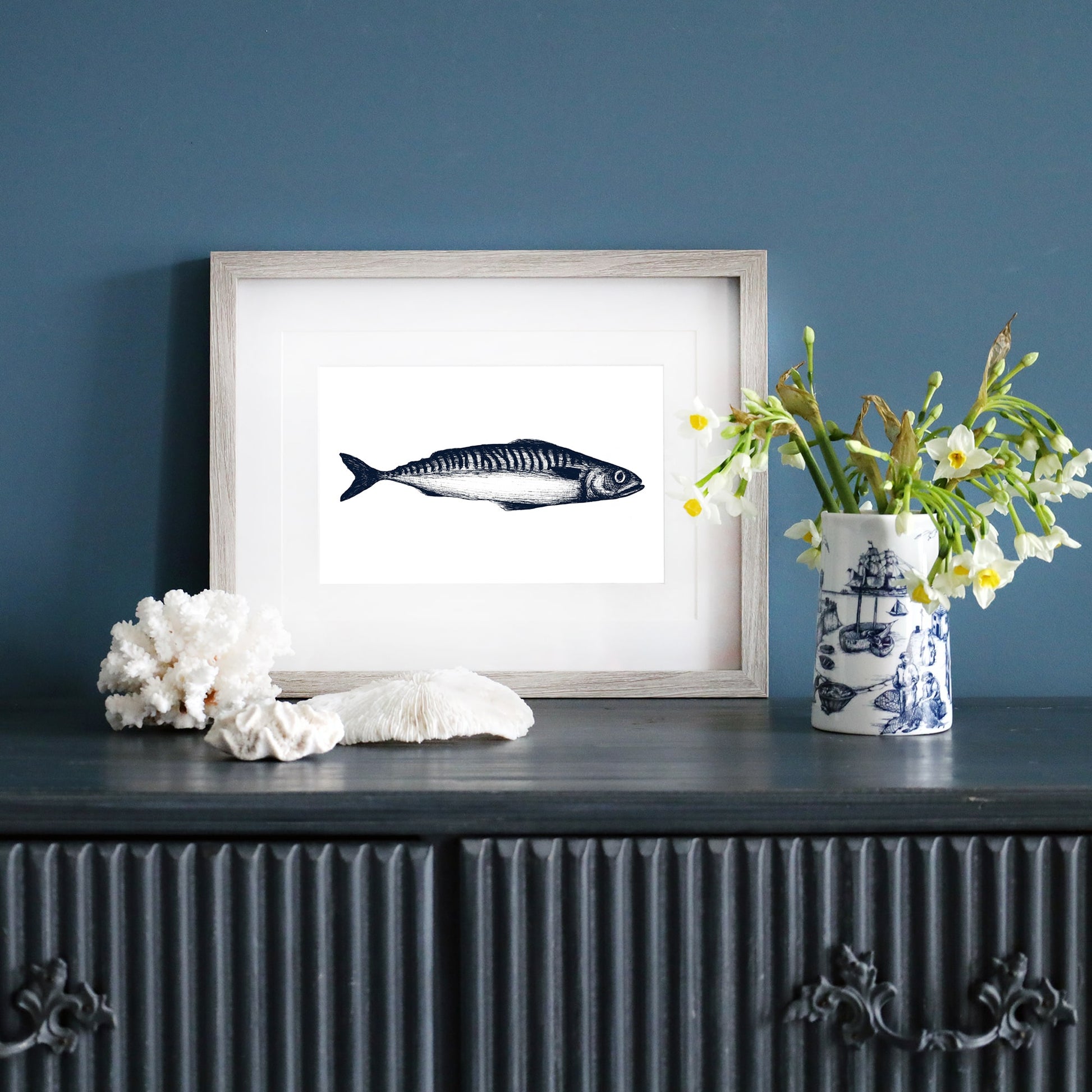 A framed blue-and-white illustration of a single mackerel in a pale wood frame, displayed on a dark blue wall. Below, a dark sideboard is decorated with white coral, a ceramic vase with yellow and white flowers, and a blue and white nautical-patterned mug.
