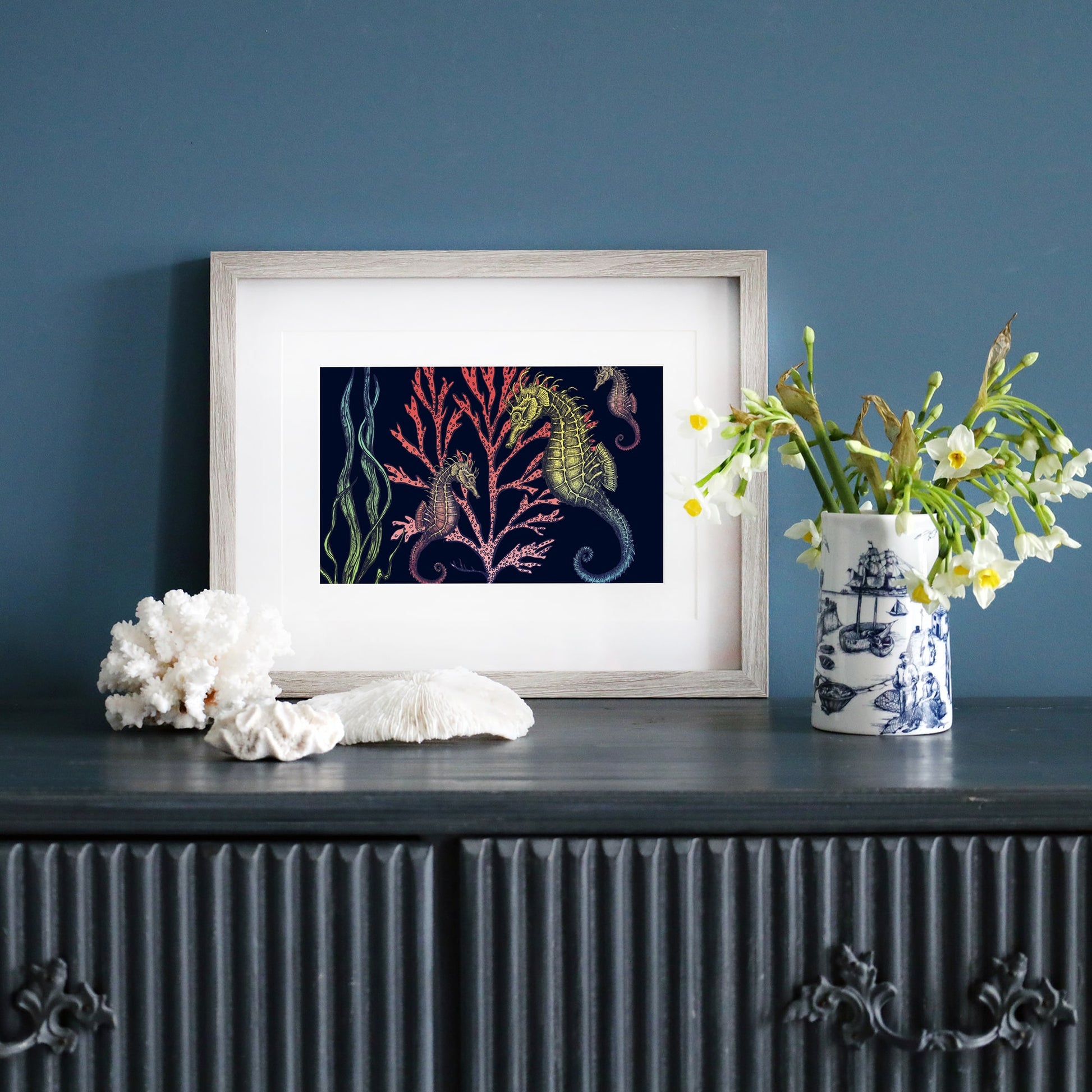 Framed print of seahorses and coral on a navy background, leaning against a deep blue wall. A dark wooden sideboard holds coral pieces, a nautical-patterned vase with flowers, and other coastal decor