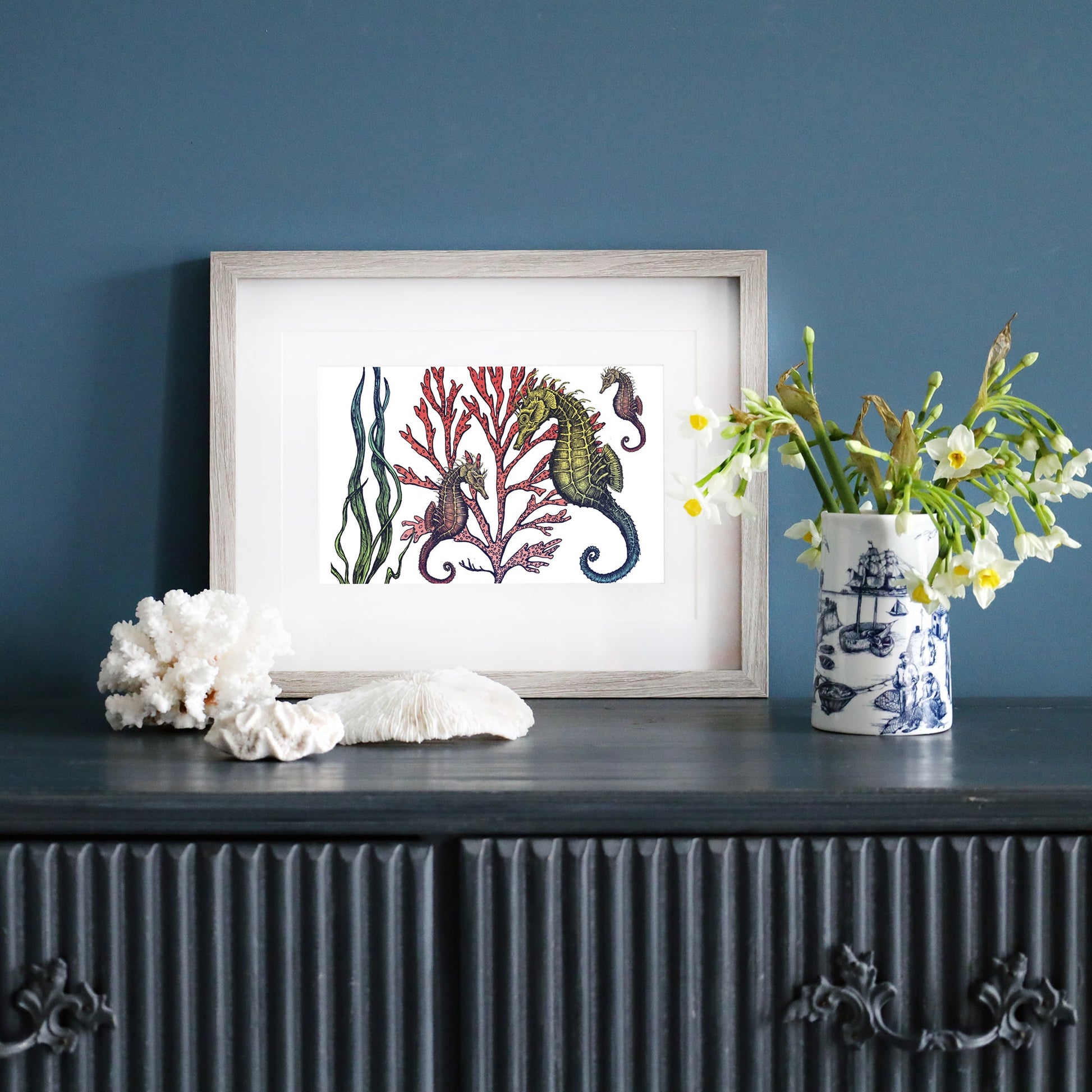 A framed illustration of seahorses among red coral and green seaweed, set on a dark blue sideboard. The frame has a light wood finish and a white mat. Next to it, a blue and white nautical-patterned vase holds fresh white flowers, with coral decorations placed on the surface.
