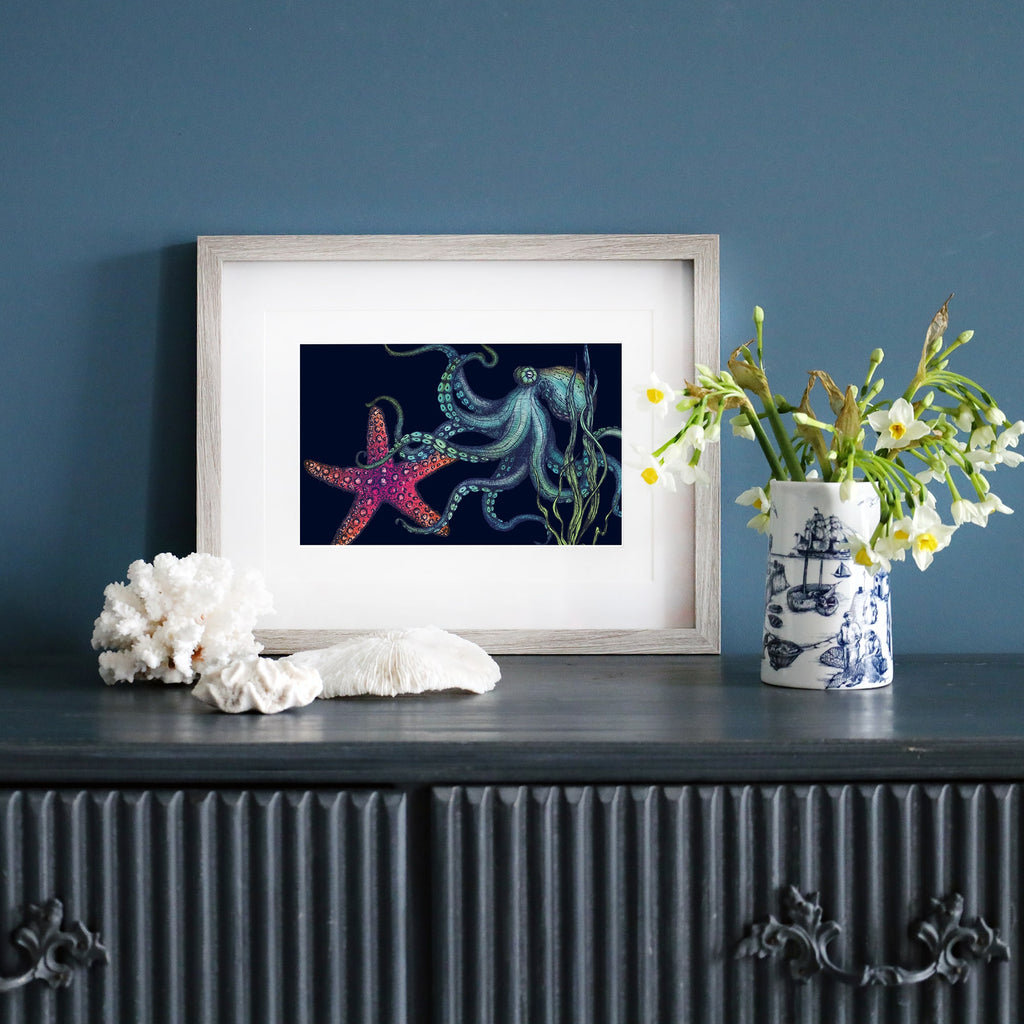 A framed illustration of an octopus and a vibrant red starfish on a deep navy background sits on a dark wooden sideboard against a blue wall. The octopus, depicted in shades of blue and green, extends its tentacles near swaying seaweed. The artwork is housed in a pale wood frame. Beside it, a blue and white nautical-themed vase holds fresh white flowers, and pieces of coral are arranged on the surface.