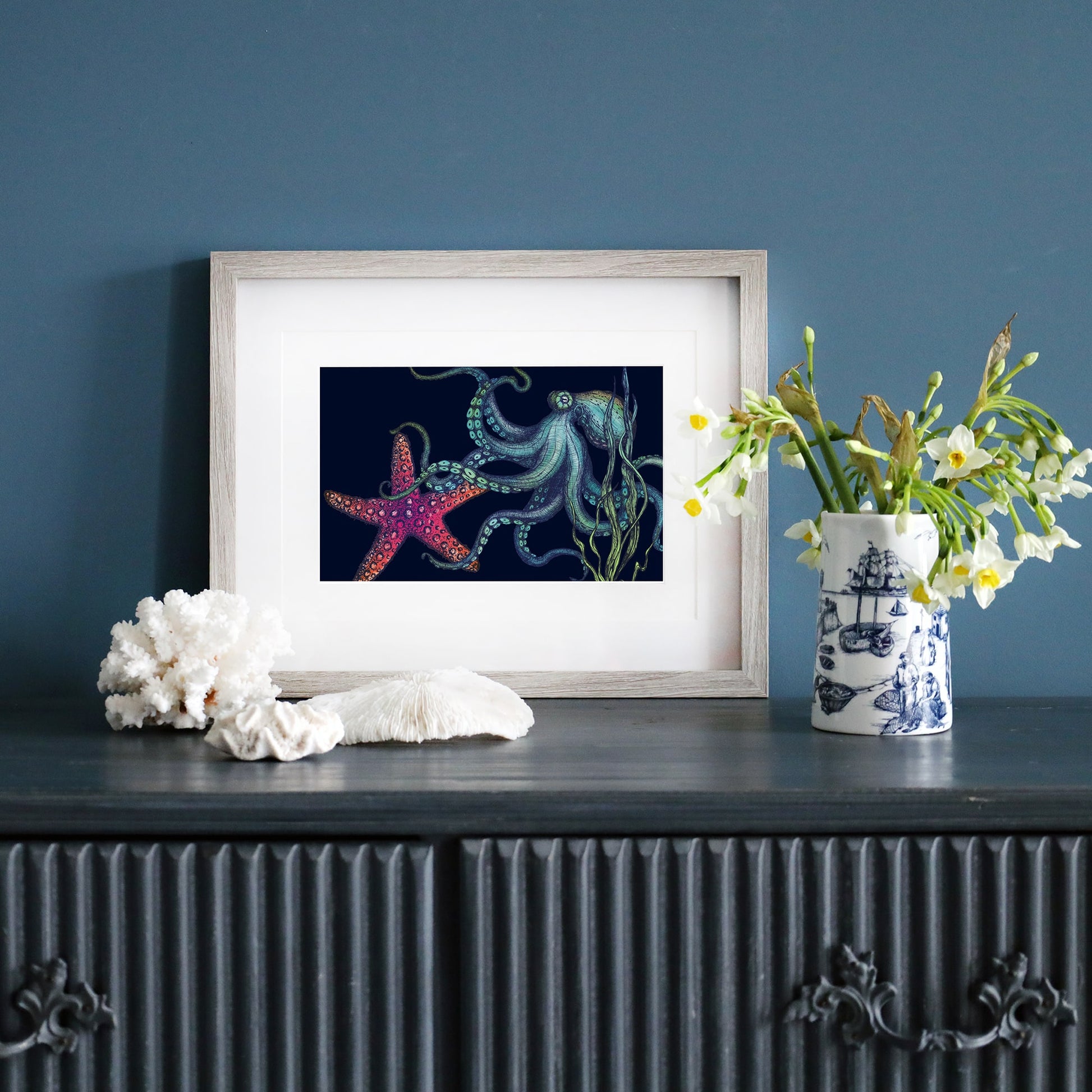 A framed illustration of an octopus and a vibrant red starfish on a deep navy background sits on a dark wooden sideboard against a blue wall. The octopus, depicted in shades of blue and green, extends its tentacles near swaying seaweed. The artwork is housed in a pale wood frame. Beside it, a blue and white nautical-themed vase holds fresh white flowers, and pieces of coral are arranged on the surface.